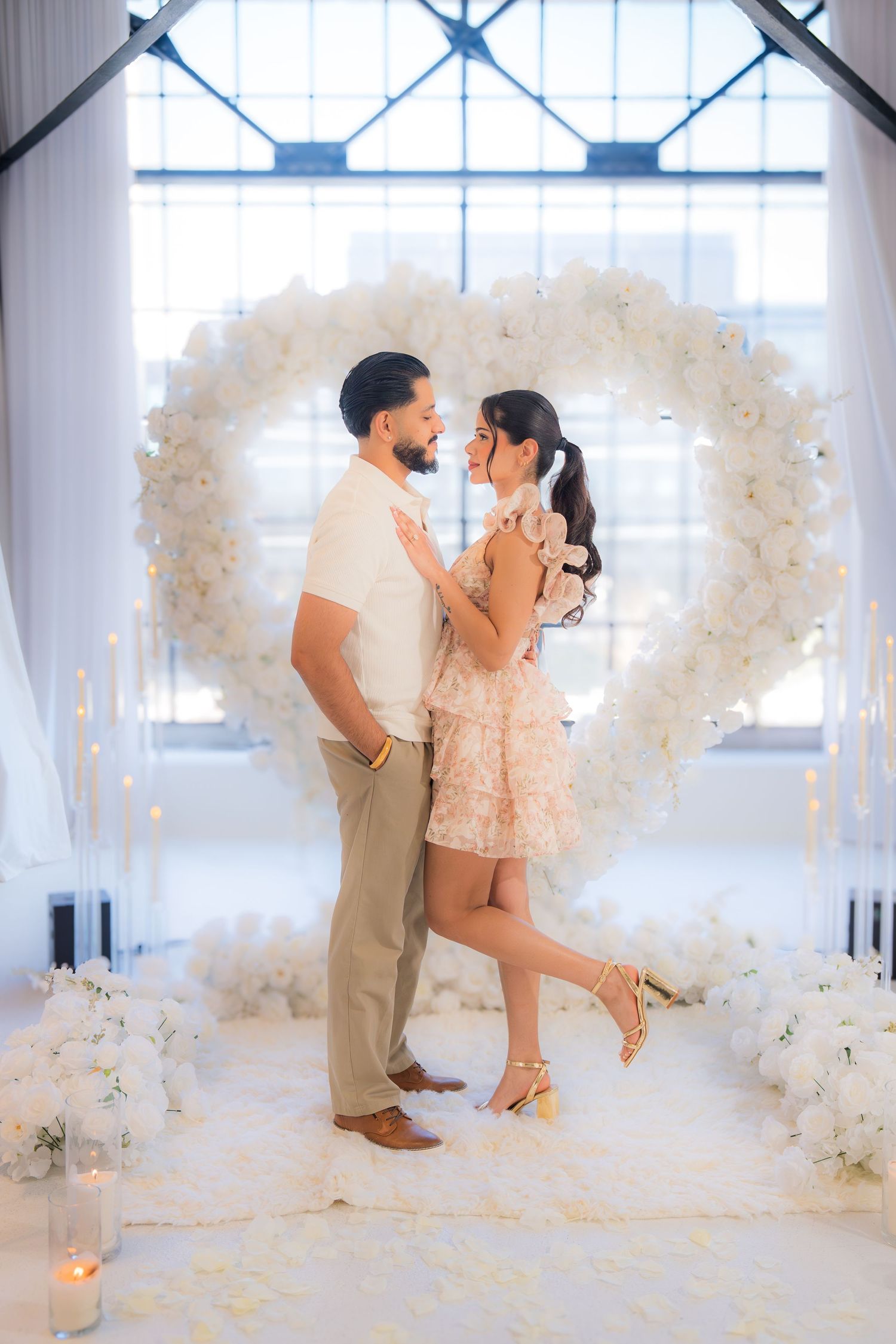 Couple embracing in front of a stunning white floral heart arch with candles and fairy lights.