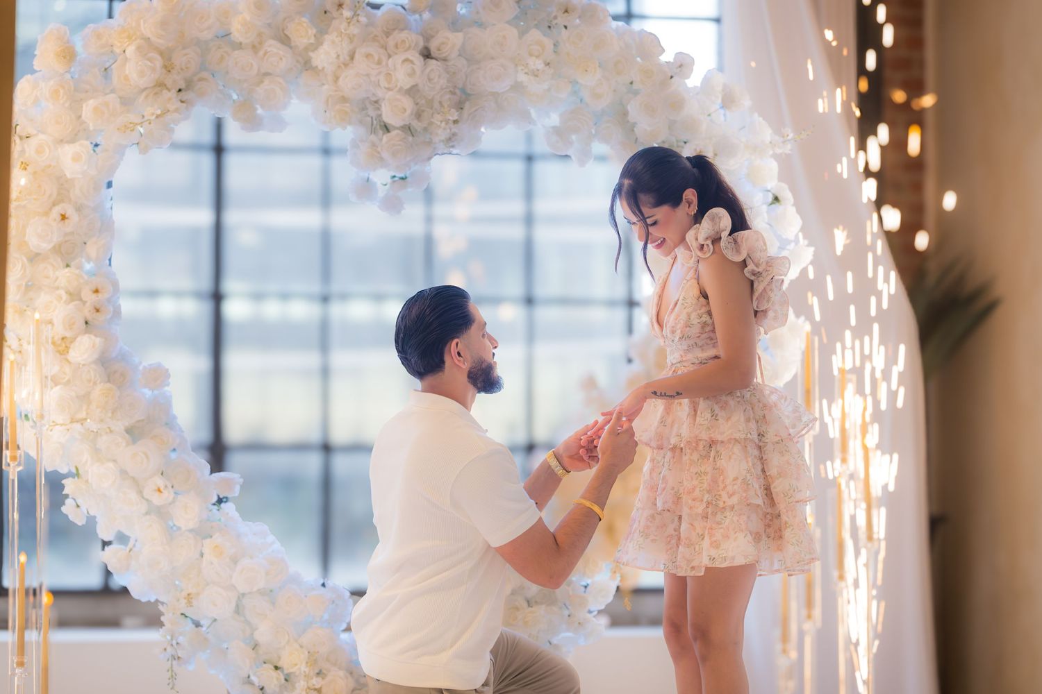 Romantic marriage proposal with man kneeling before his partner under a glowing white floral heart arch.