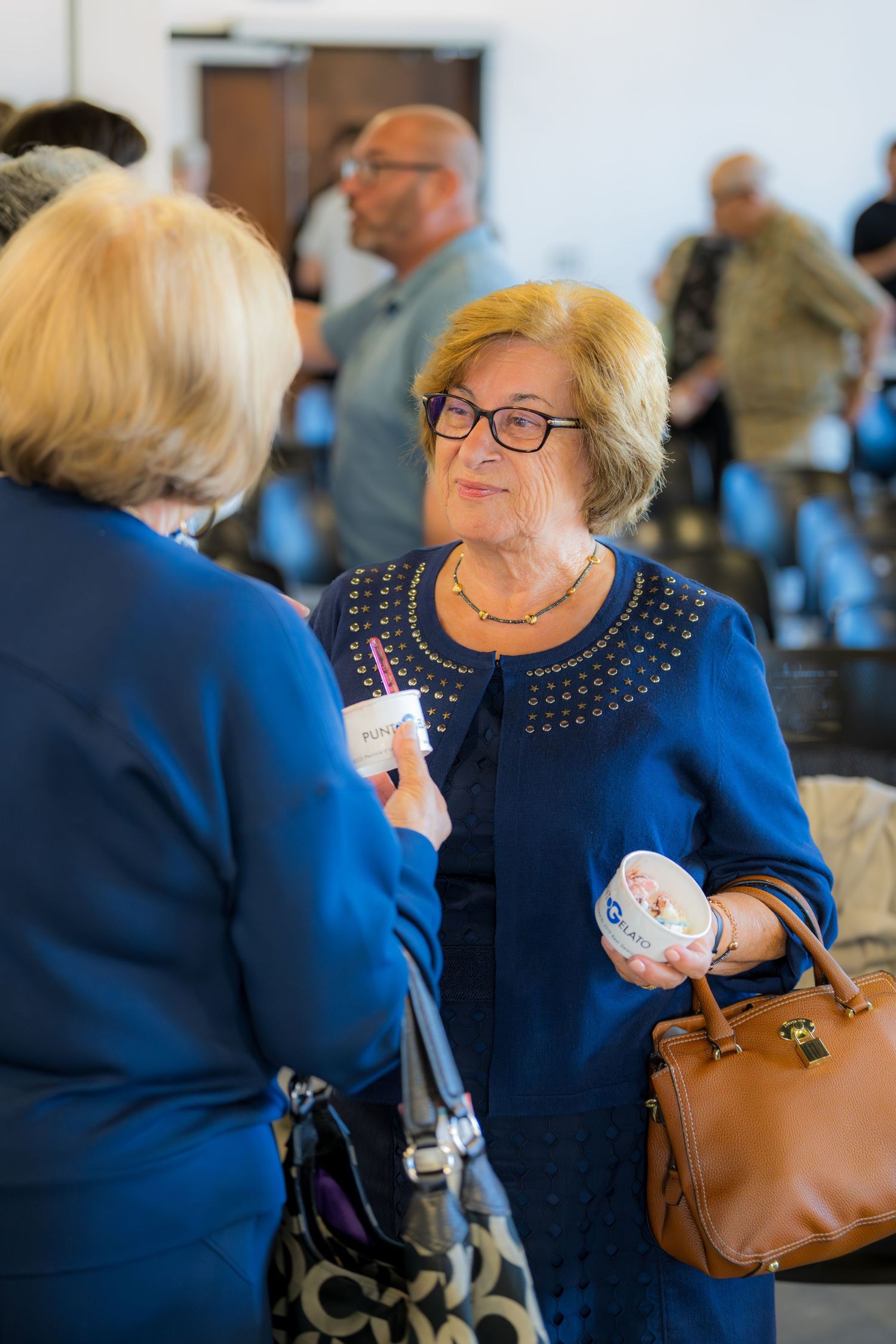 Two senior women in blue outfits conversing at a social gathering, one holding a decorative bowl and purse.