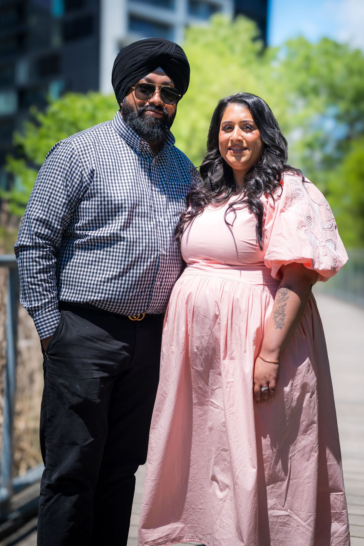 A Sikh man in a black turban and checkered shirt poses with a woman in a pink dress outdoors.
