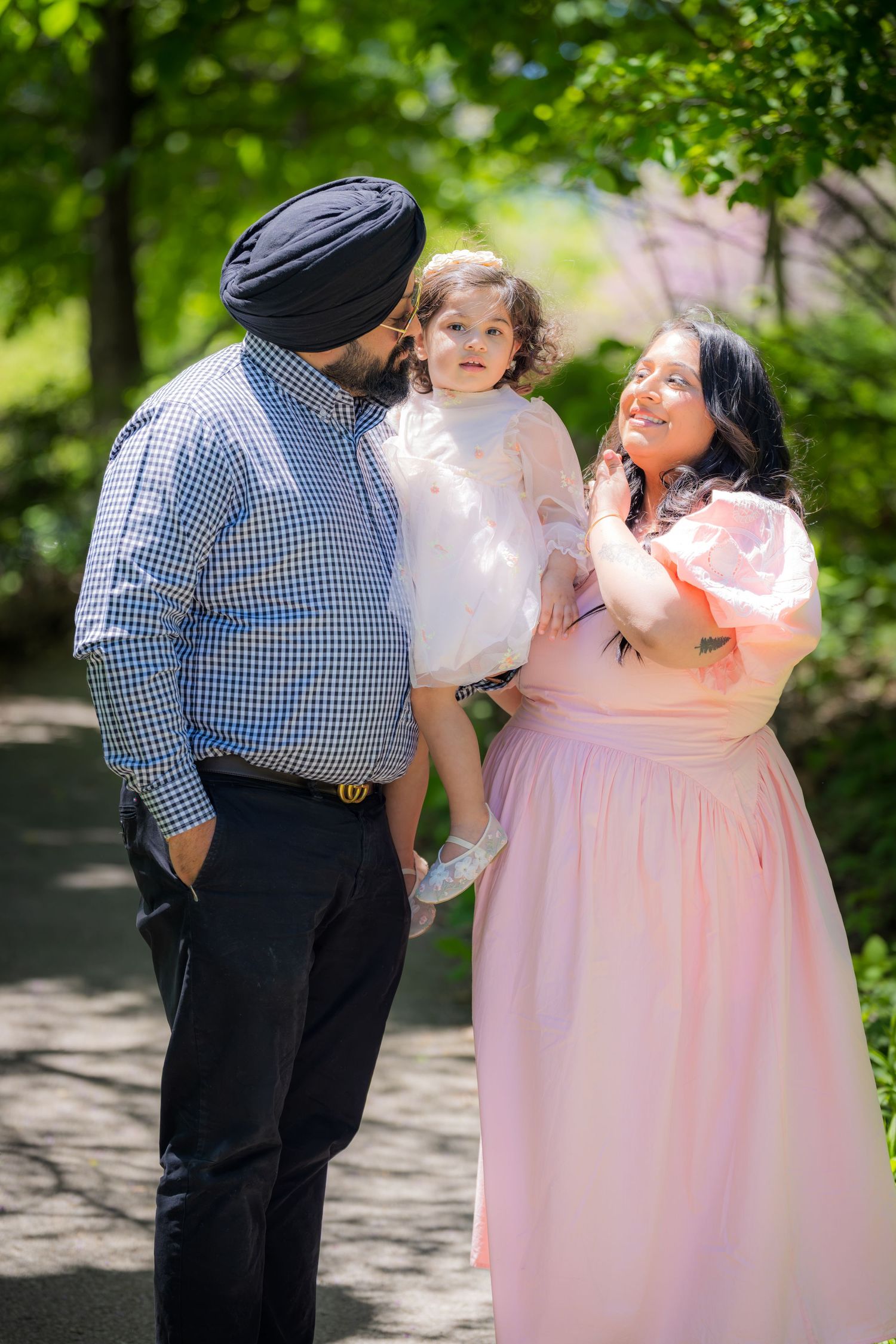 Sikh father in turban and mother in pink dress lovingly holding toddler daughter outdoors.