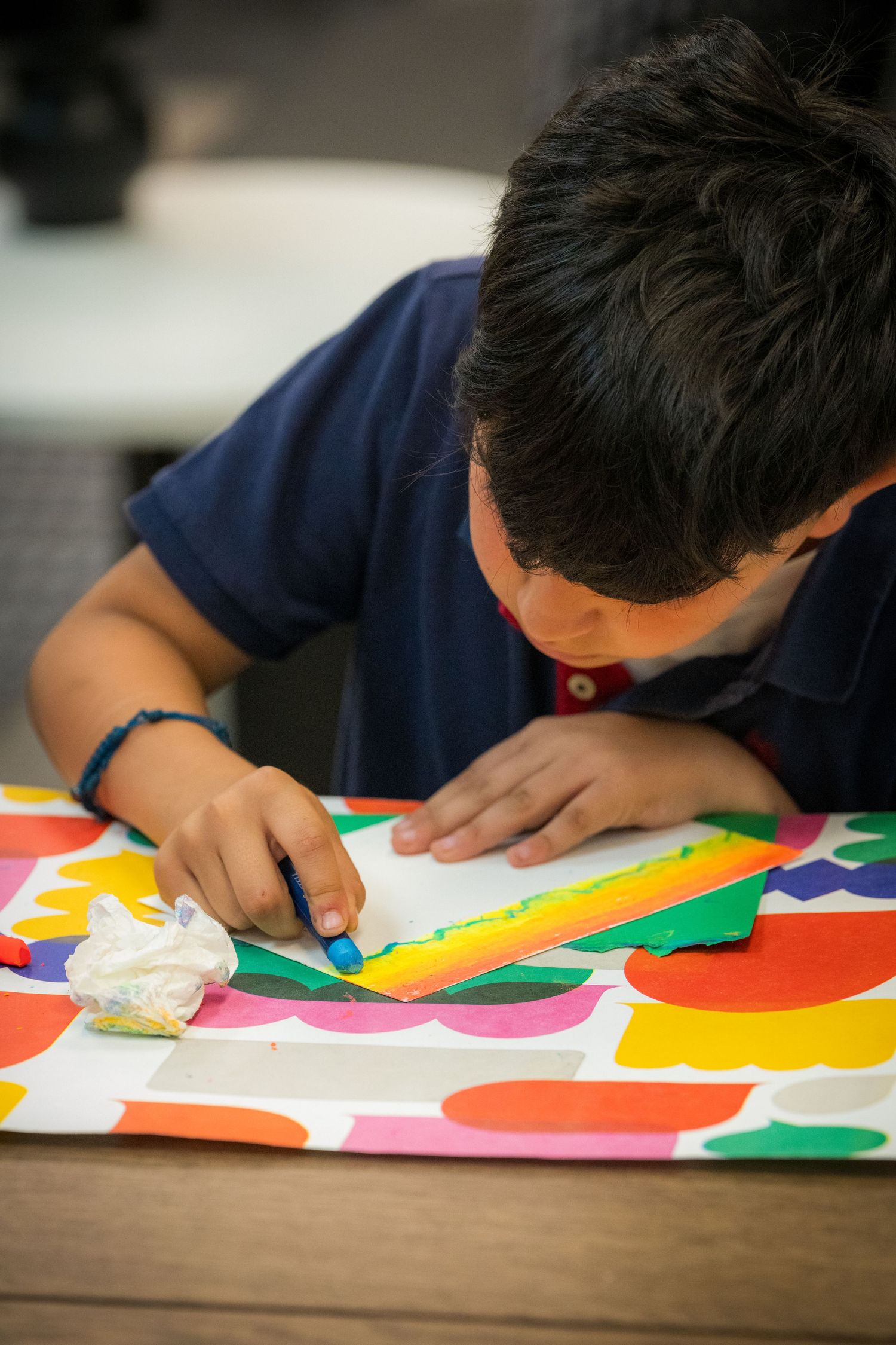 Young boy concentrating on a colorful art project using a blue crayon and ruler on a bright patterned surface.