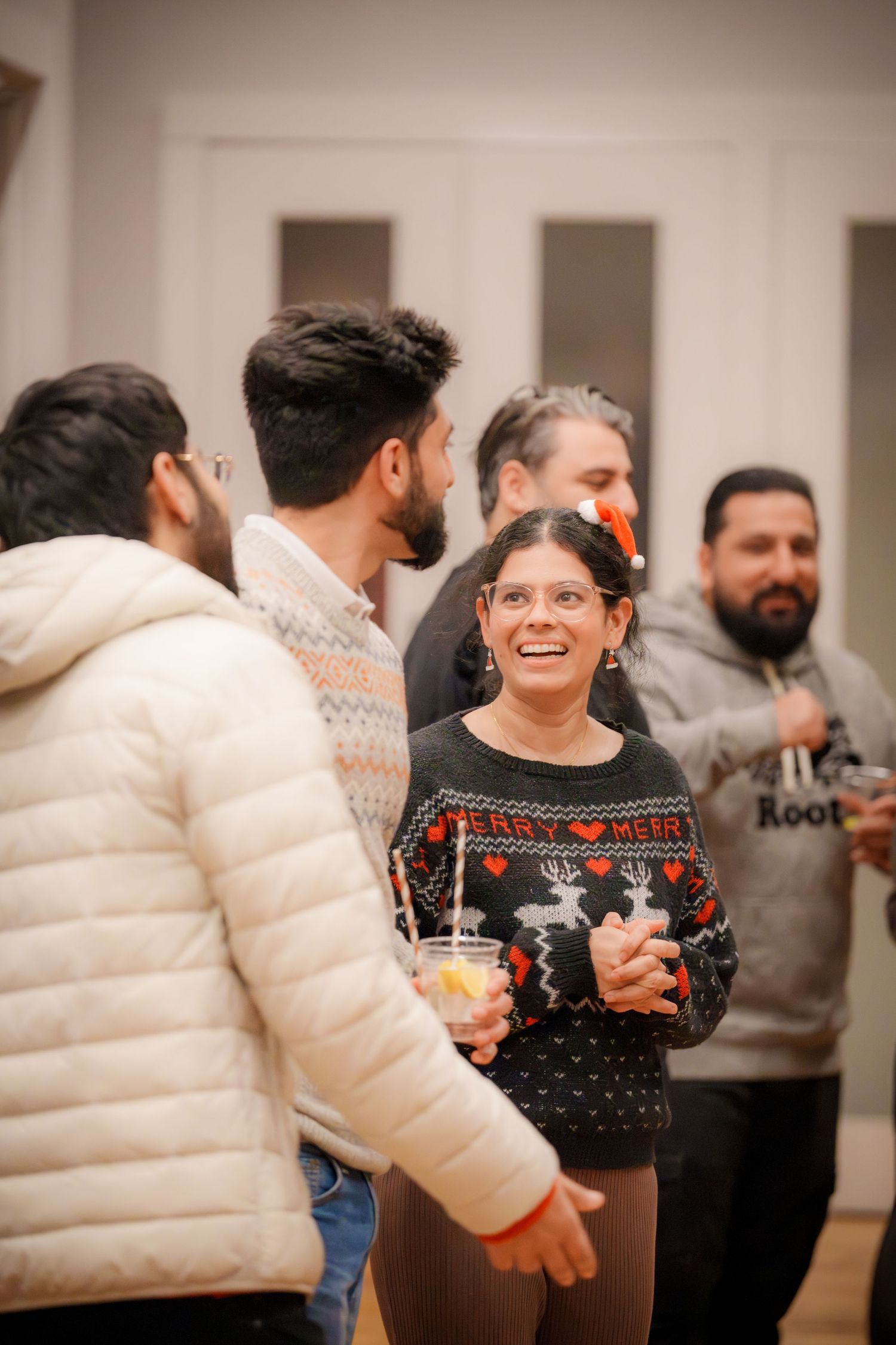 Group wearing holiday sweaters and winter coats socializing at an indoor party.
