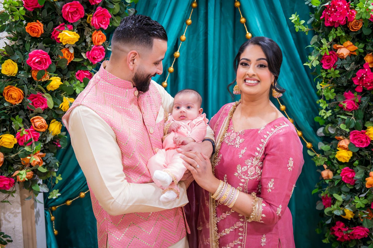 South Asian couple in pink traditional attire holding their baby at a floral decorated ceremony backdrop.