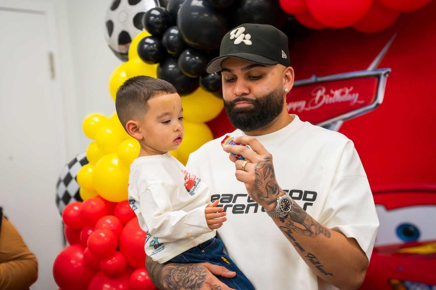 Man holding toddler at a Cars-themed birthday party with red, yellow, and black balloon decorations.