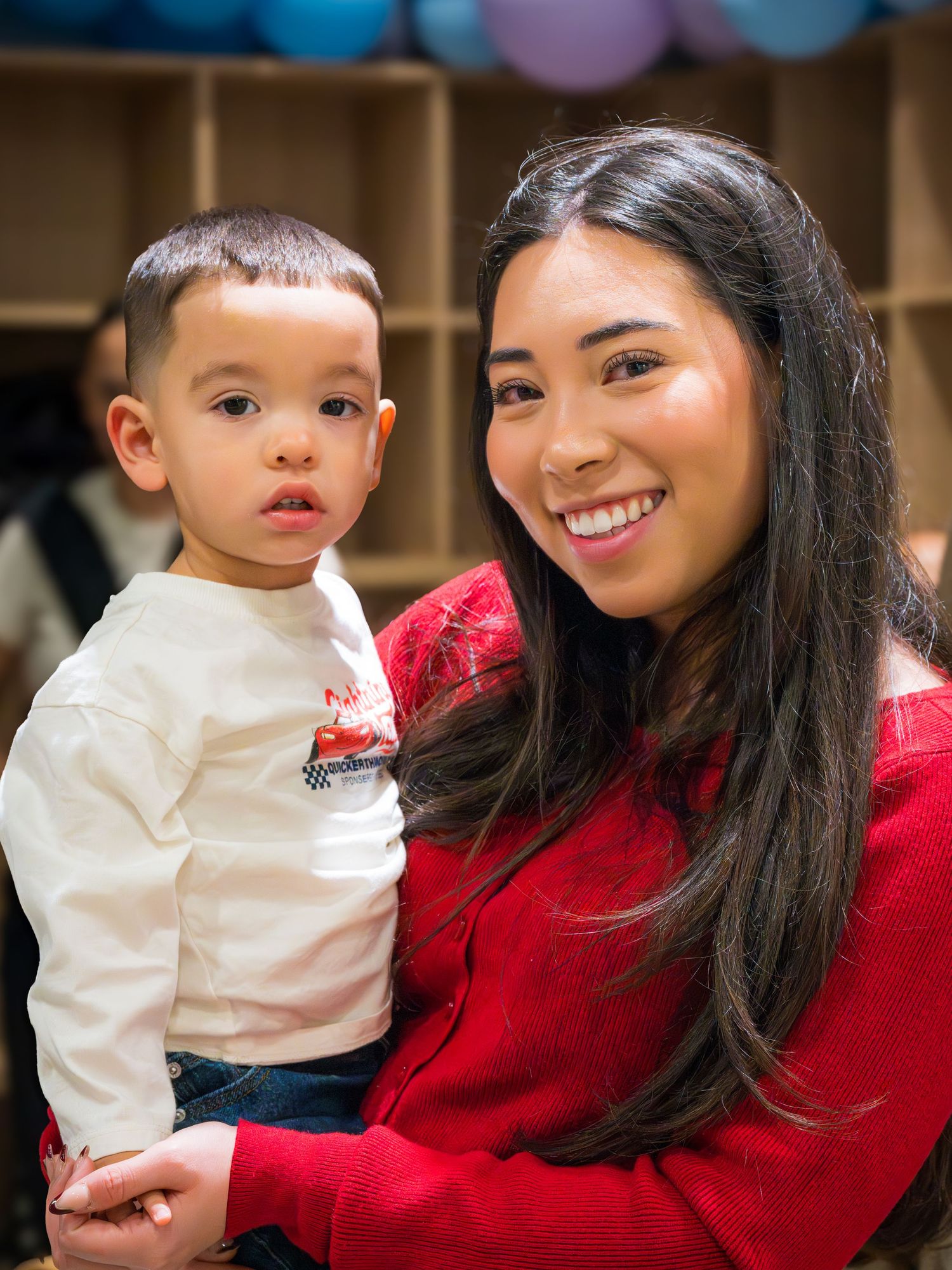 Smiling young woman in red sweater holding a toddler boy in a Lightning McQueen shirt at an indoor event.