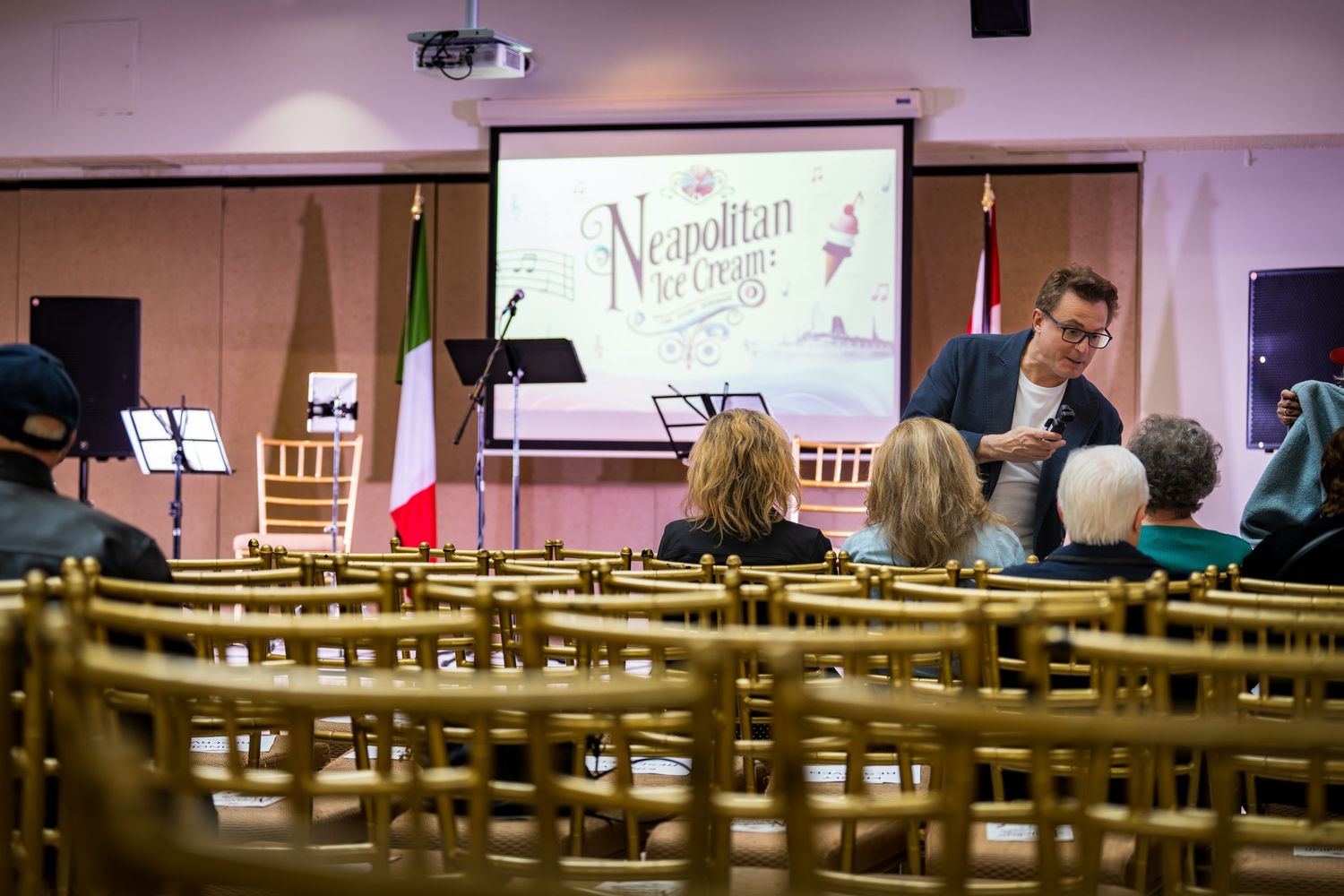 A speaker presents at a Neapolitan heritage event in a decorated hall with Italian flag and seated audience.