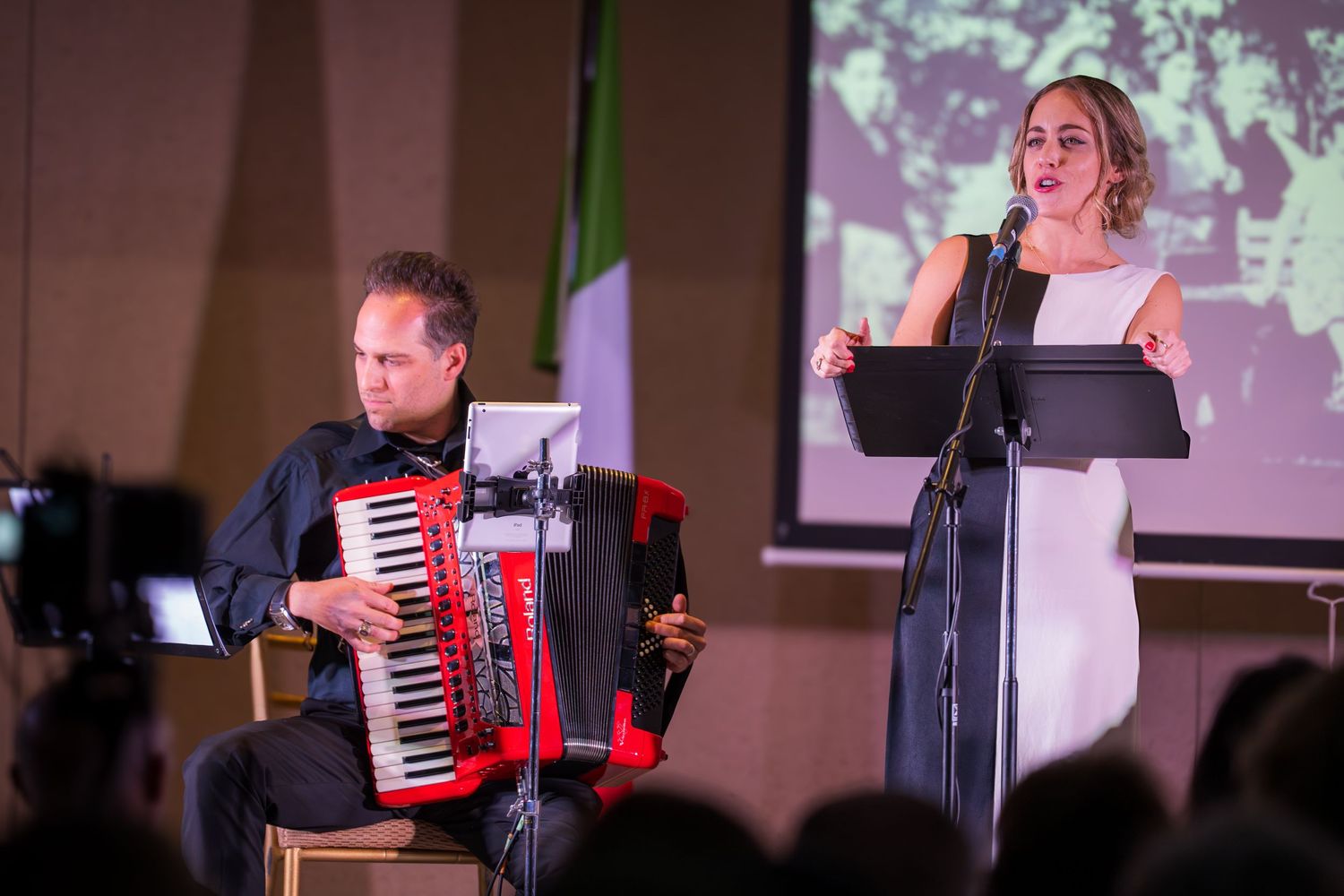 Male accordionist with red Roland accordion performs alongside female singer in black and white dress on stage.