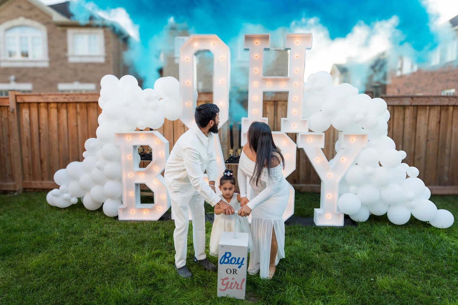 A couple and toddler stand before an OH BABY marquee sign with white balloons at a gender reveal party.