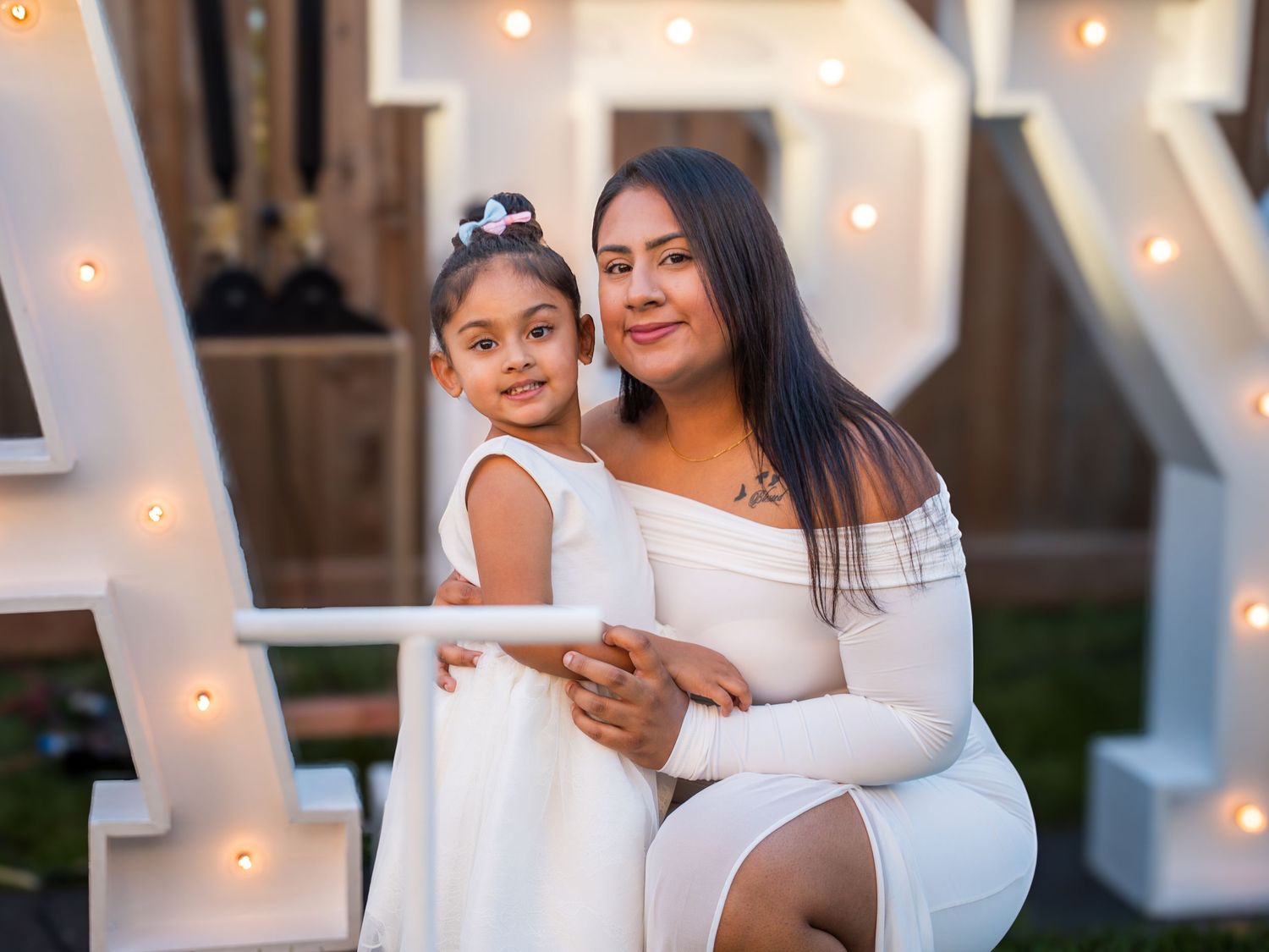 A woman and young girl in matching white dresses pose together in front of illuminated marquee letters at a celebration.