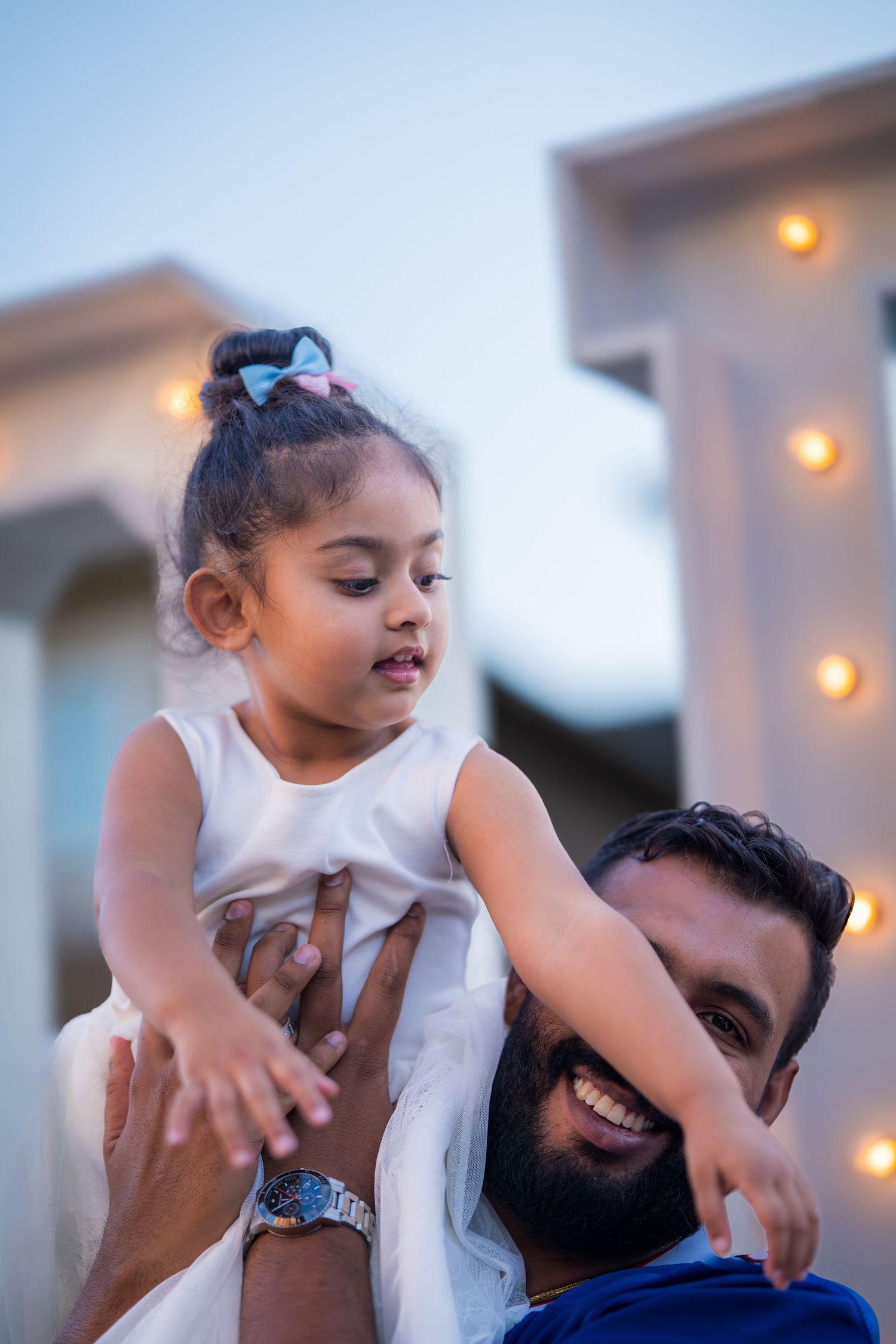 Joyful bearded father lifting young daughter in white dress against twilight sky at outdoor celebration.