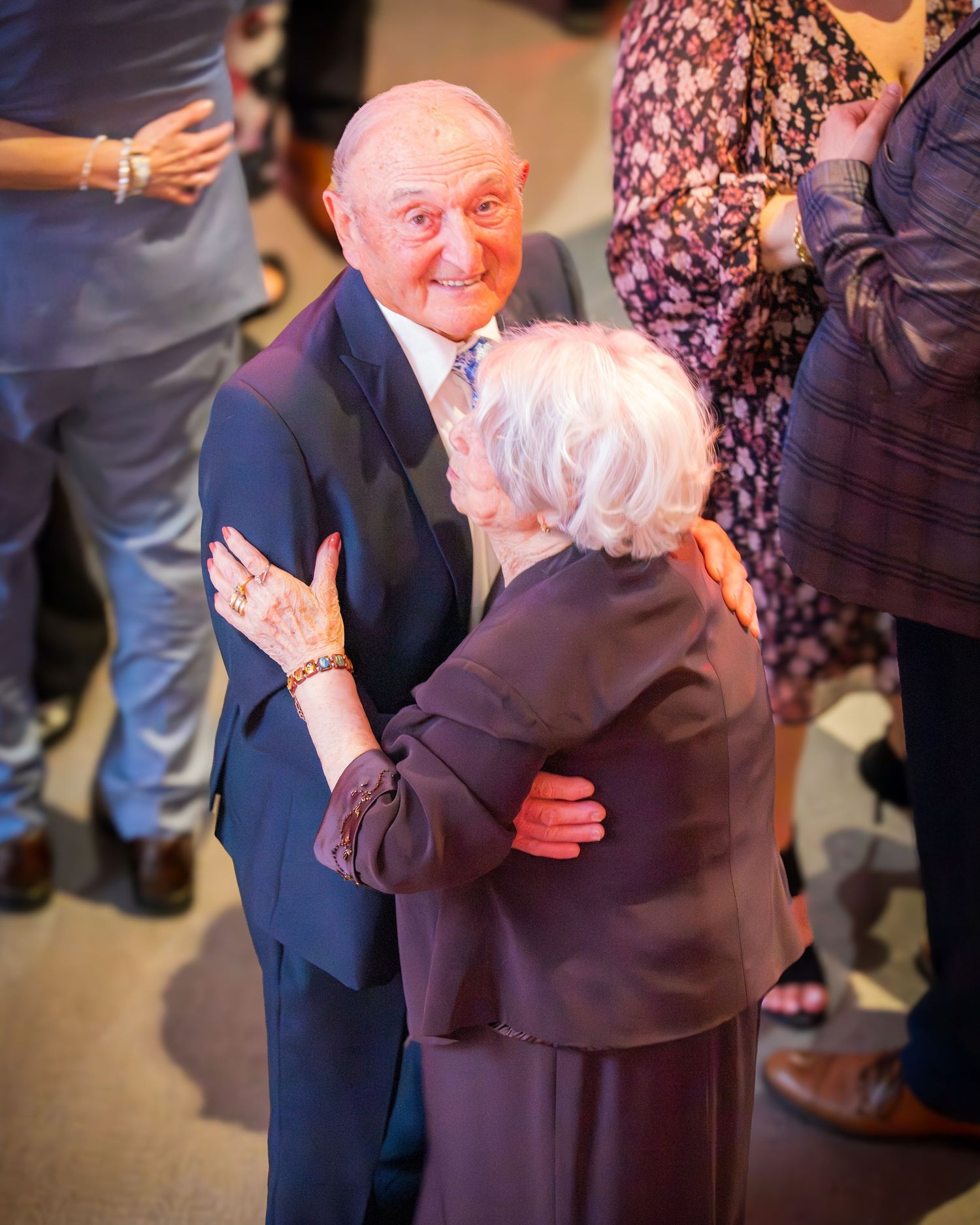 Elderly man in navy suit warmly embraces an older woman with white hair at a formal event.