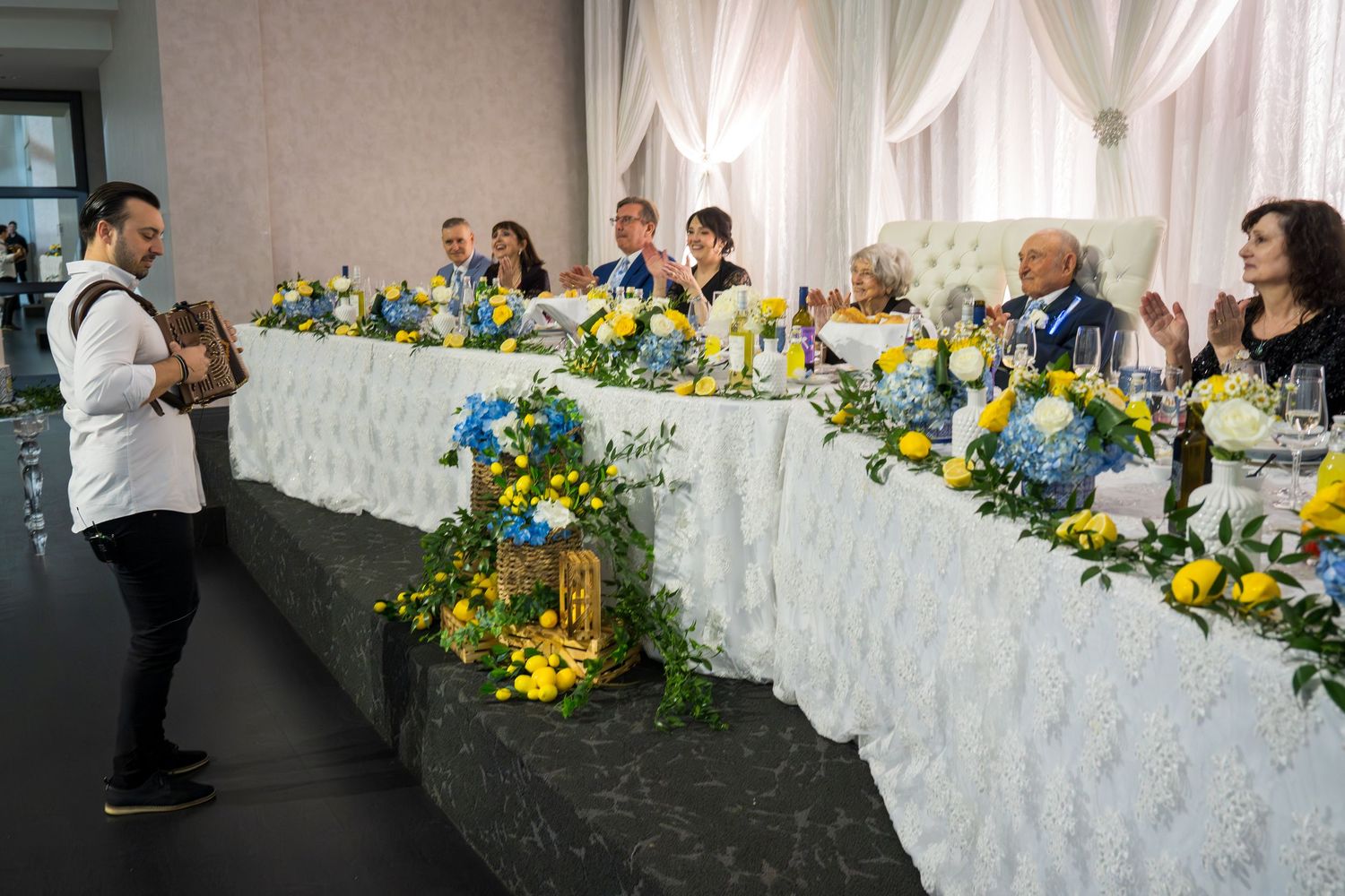 Guests applaud at an elegant wedding reception with blue and yellow floral arrangements at a decorated head table.