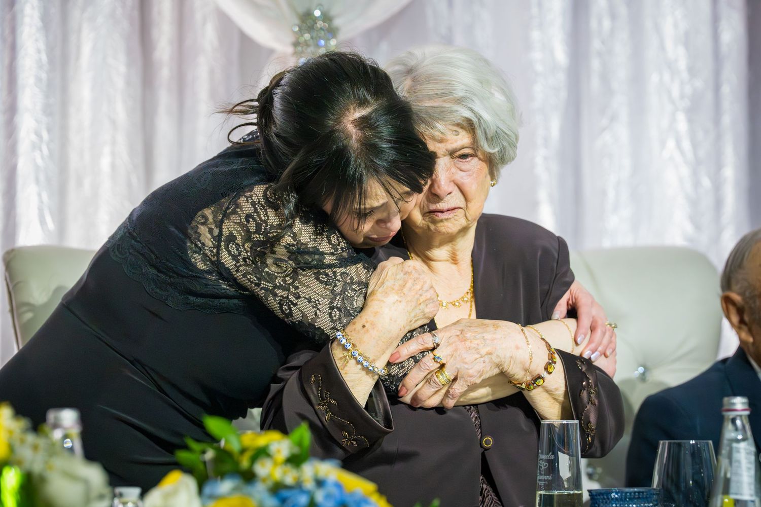 Woman embracing elderly seated woman in tender moment during anniversary celebration party reception.