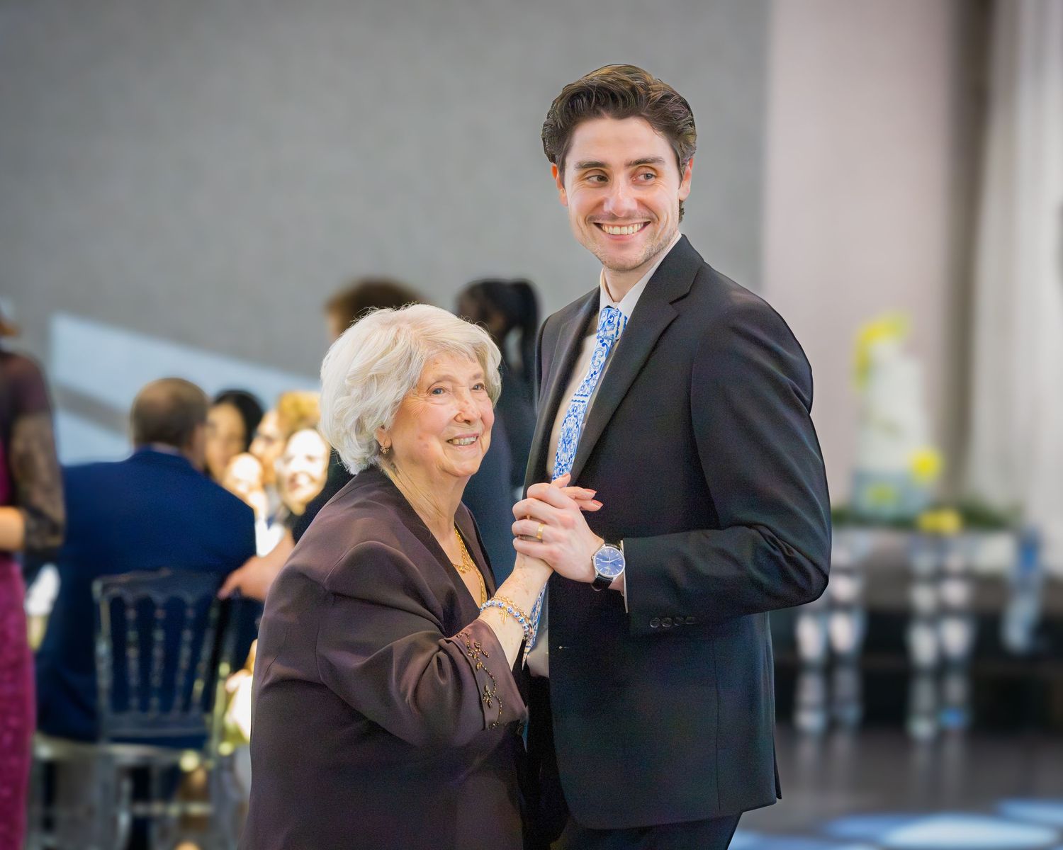 Young man in dark suit dancing with elderly woman in brown jacket at a wedding reception.