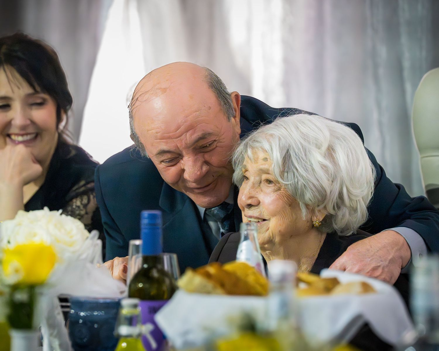 Elderly man affectionately leaning toward white-haired woman at a wedding reception dinner table.