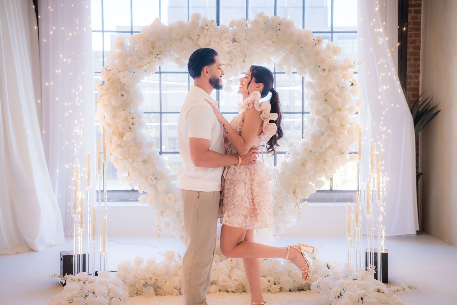 Romantic couple sharing a kiss under a white flower heart arch with fairy lights.