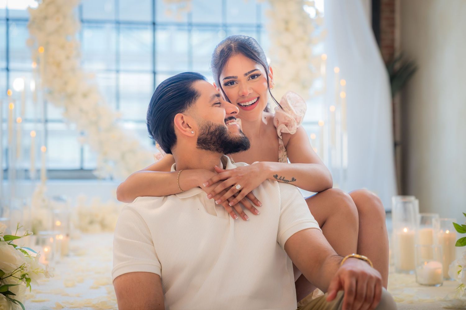 Newly engaged couple embracing and smiling surrounded by candles and white floral arrangements.