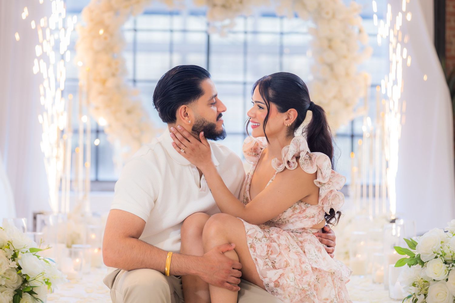 Couple sitting together and smiling at each other in front of a romantic white floral heart arch with fairy lights.
