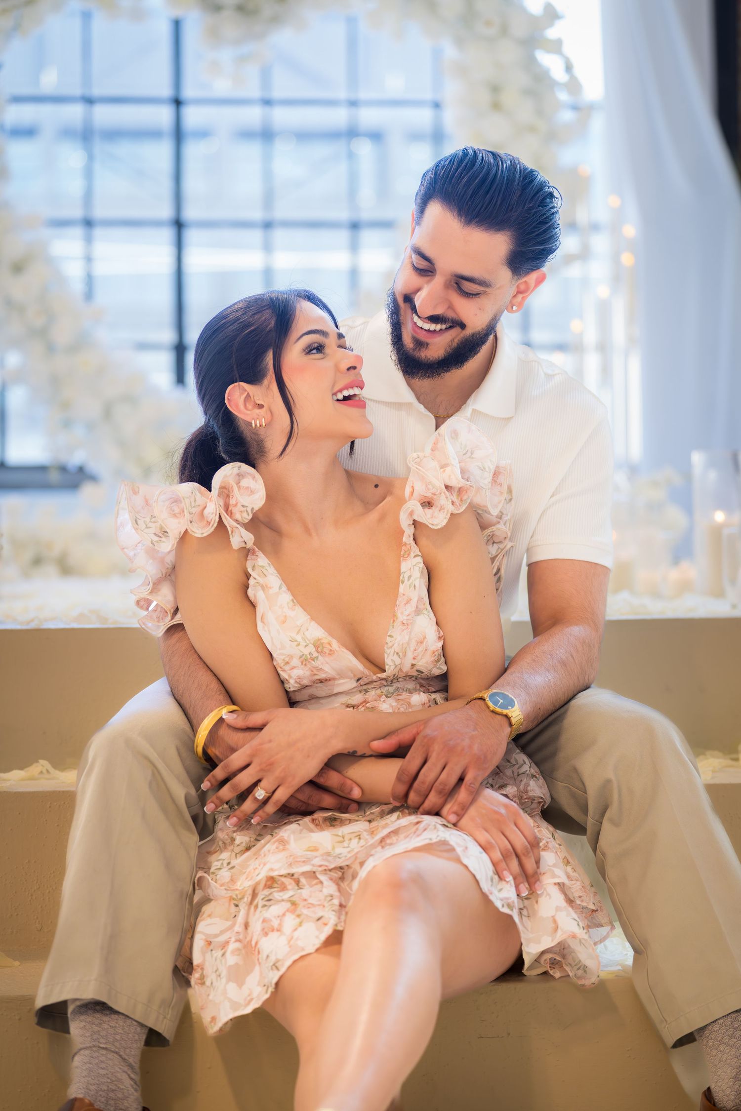Happy couple sitting closely together laughing at romantic white floral engagement celebration.