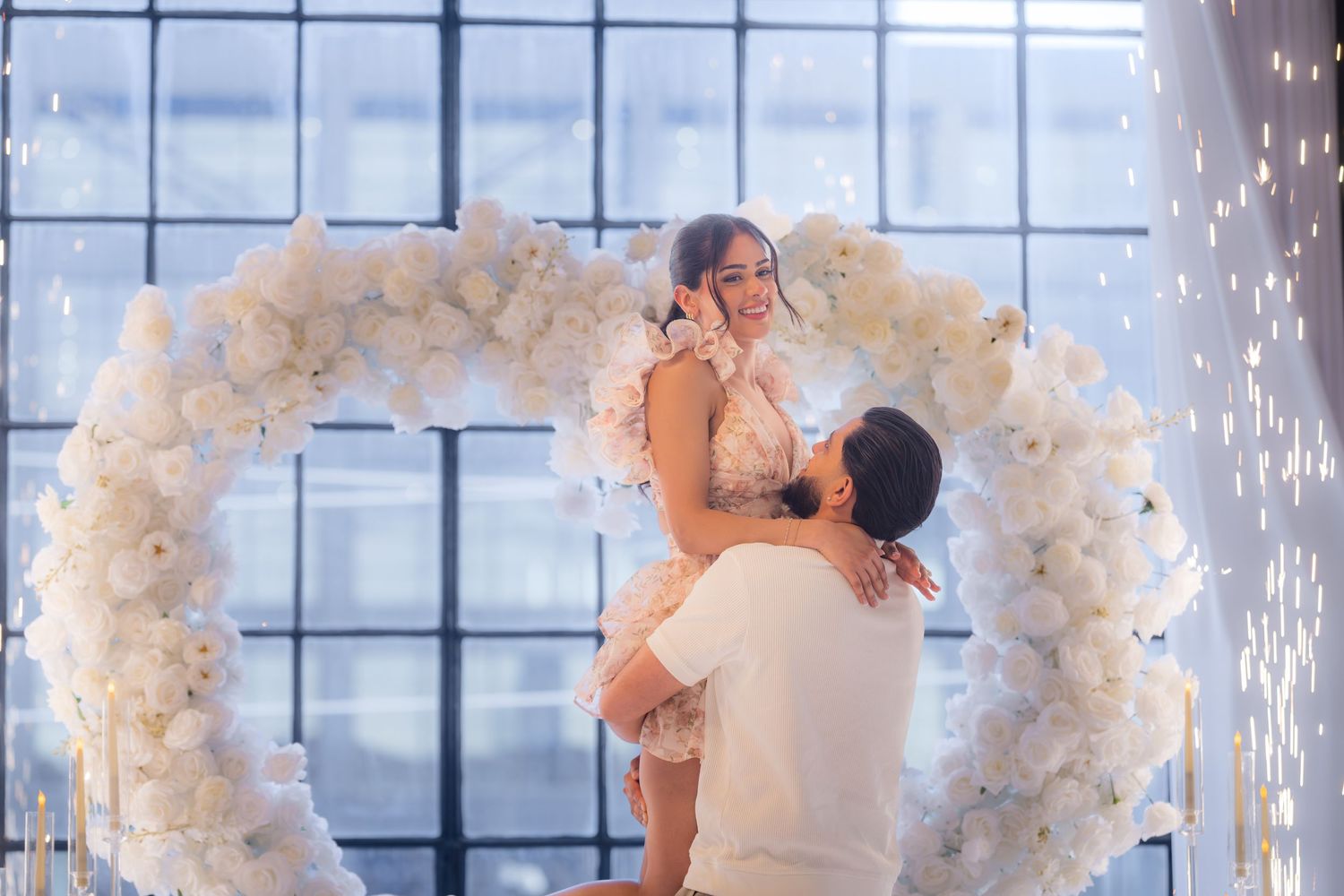 Couple embracing under a white floral heart arch with sparkling lights during a romantic proposal moment.