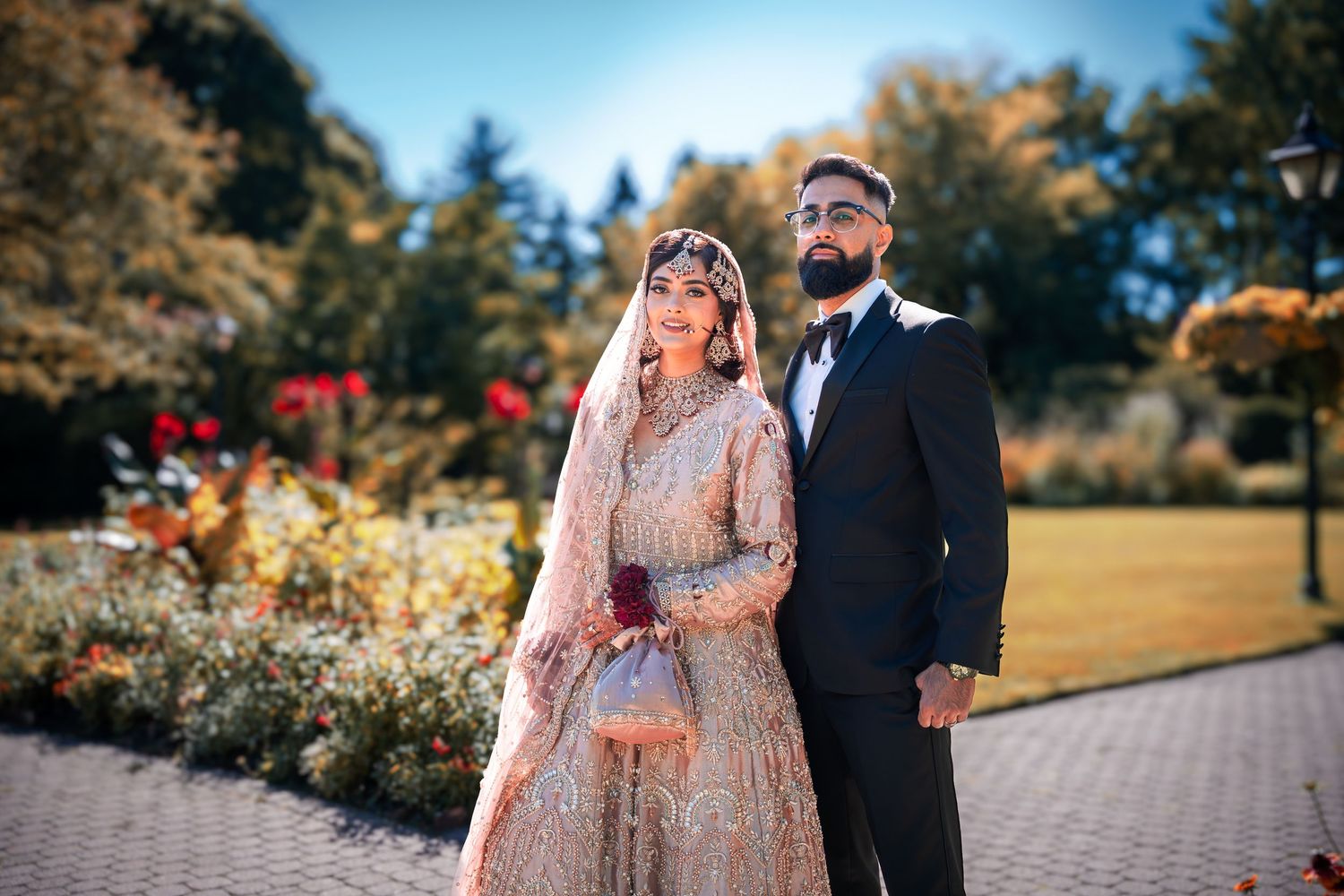 A couple stands together in a garden wearing traditional South Asian wedding attire against colorful flowers.