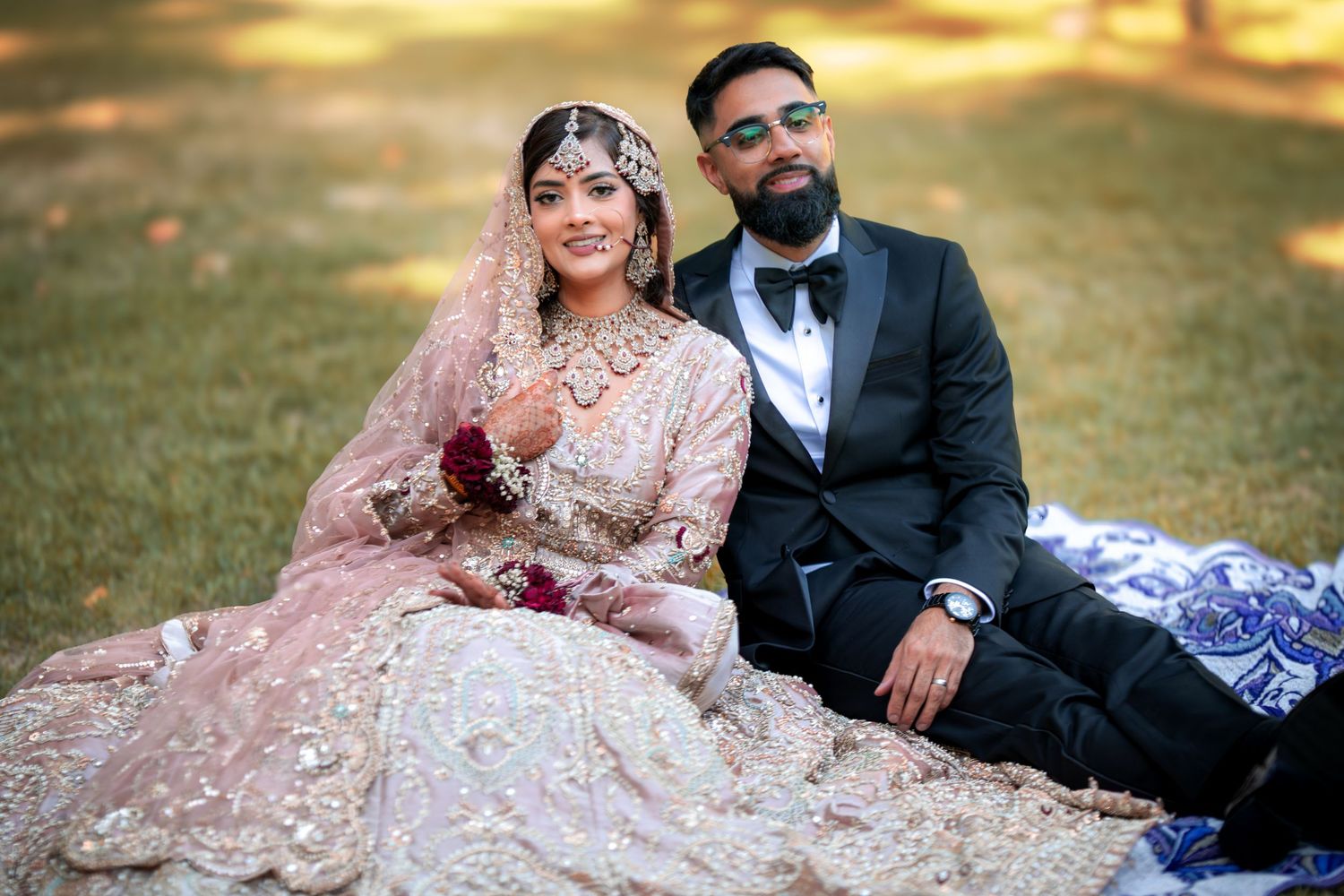 Wedding couple sits together on grass during golden hour in formal South Asian wedding attire.