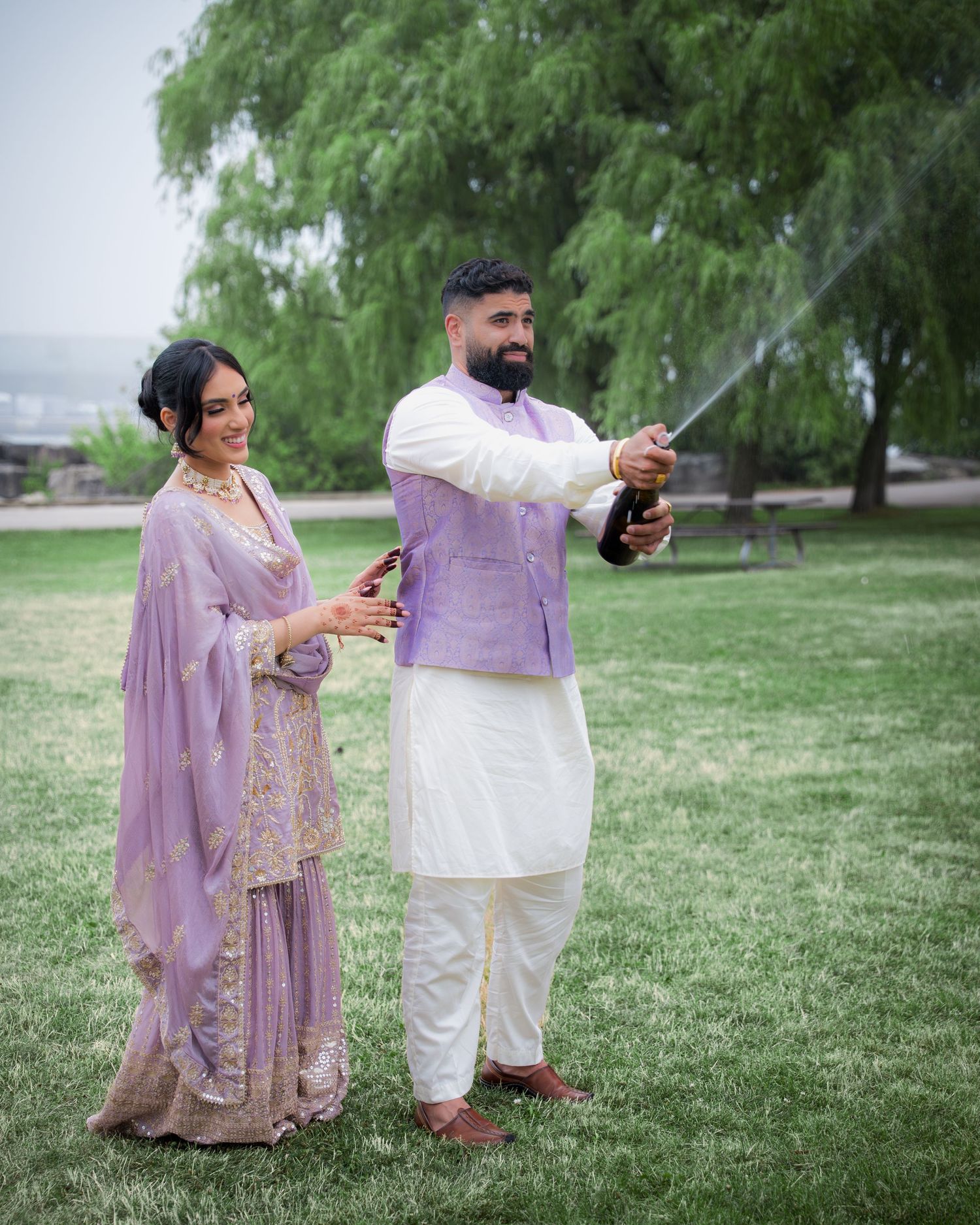 Couple in traditional Indian dress poses casually on grass with phones in hand.