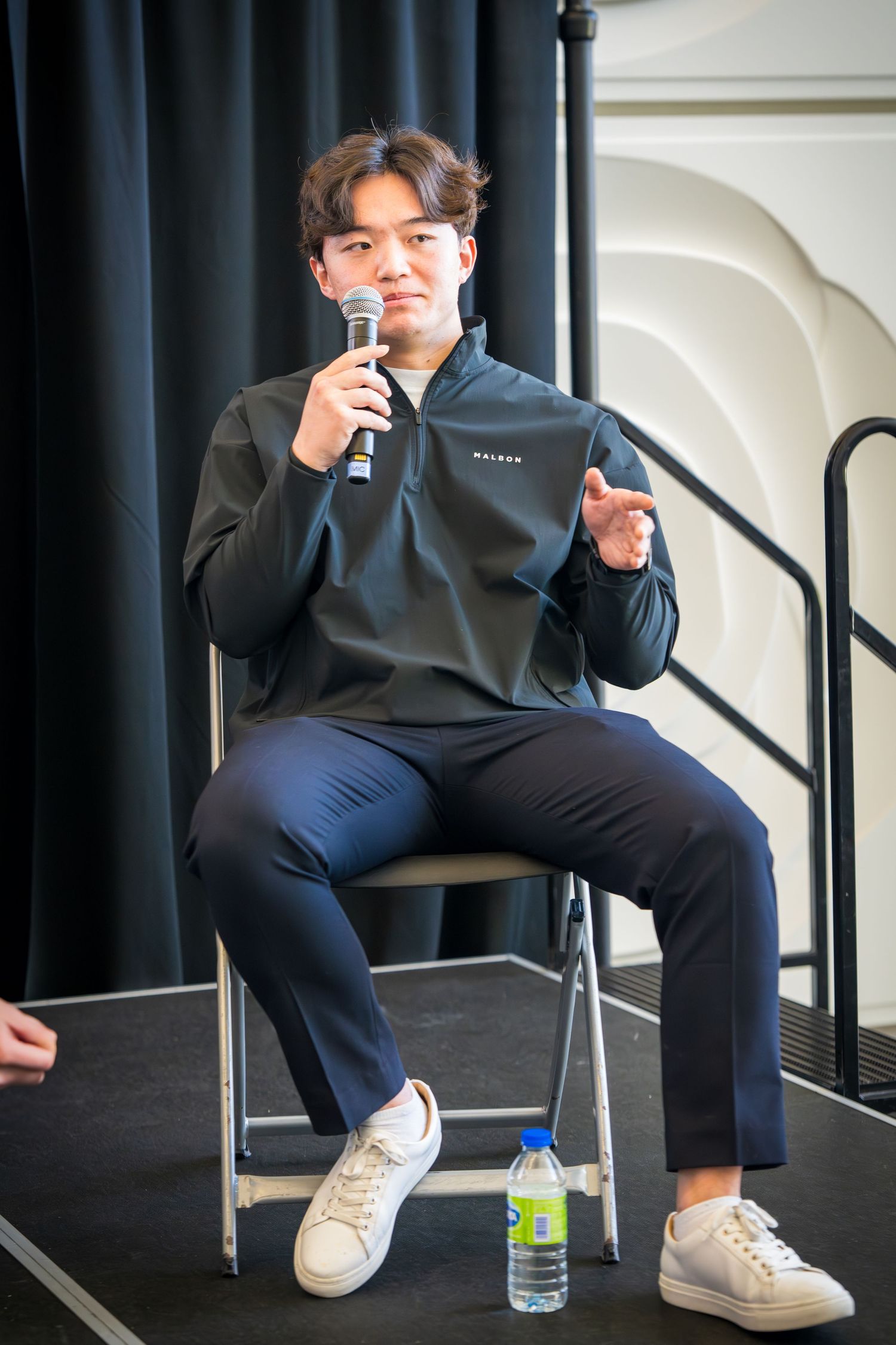 Speaker in dark attire seated on folding chair with water bottle during panel discussion.