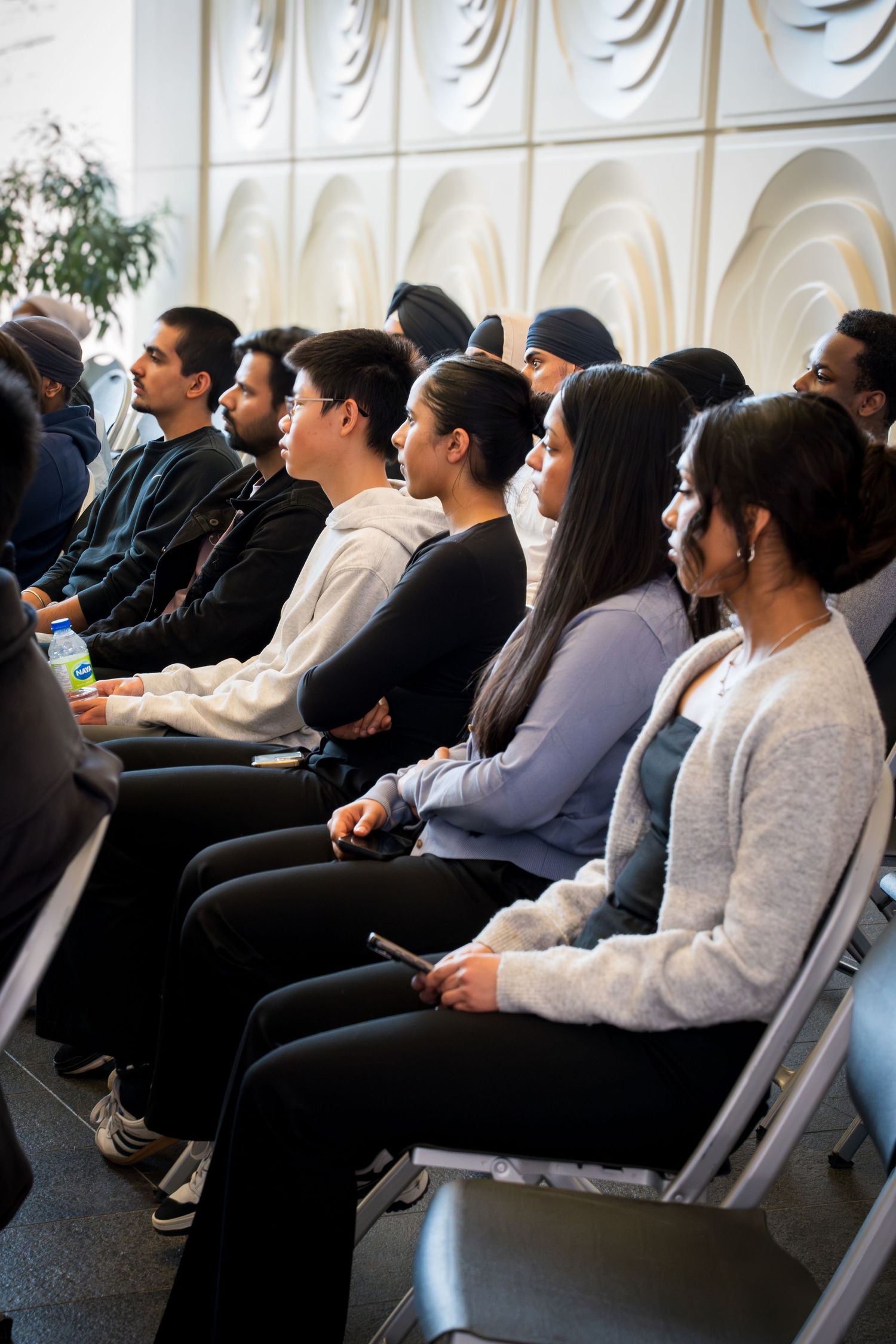 Audience members seated in folding chairs watching an educational presentation in a well-lit venue.