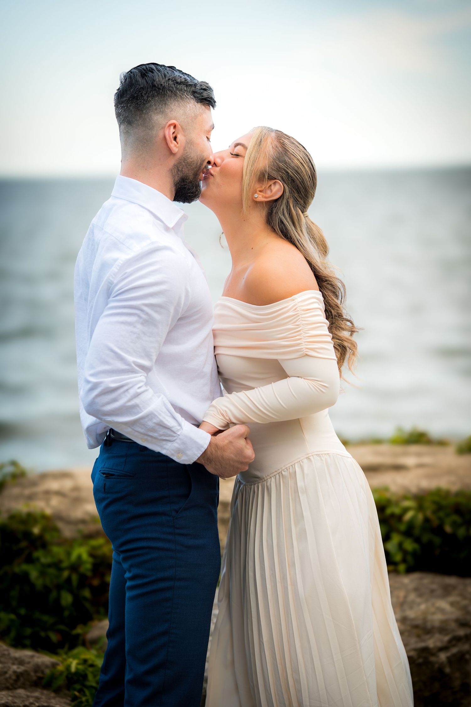 Romantic couple embracing outdoors, with bride wearing off-shoulder cream dress and groom in white shirt and navy pants.