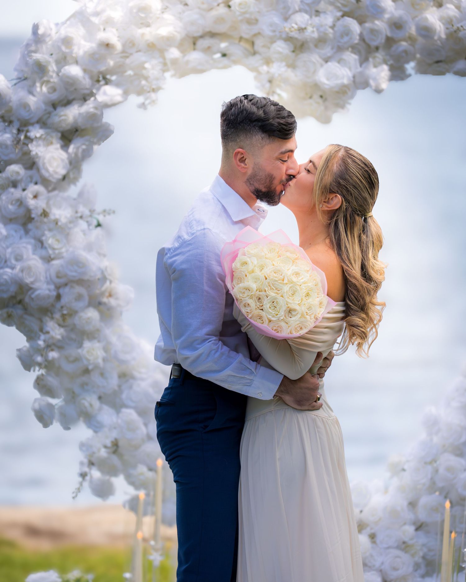 Romantic wedding moment captured under luxurious white floral arch with ocean view.