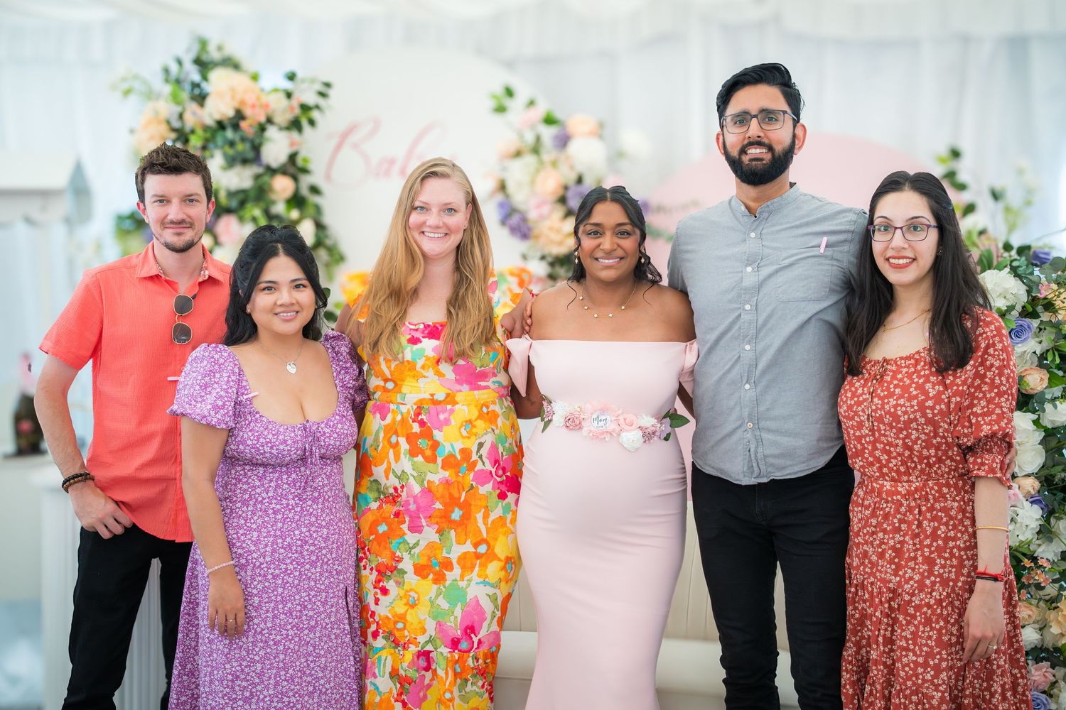 Friends gathered at a baby celebration wearing coordinated pink and floral dresses against a decorated backdrop.