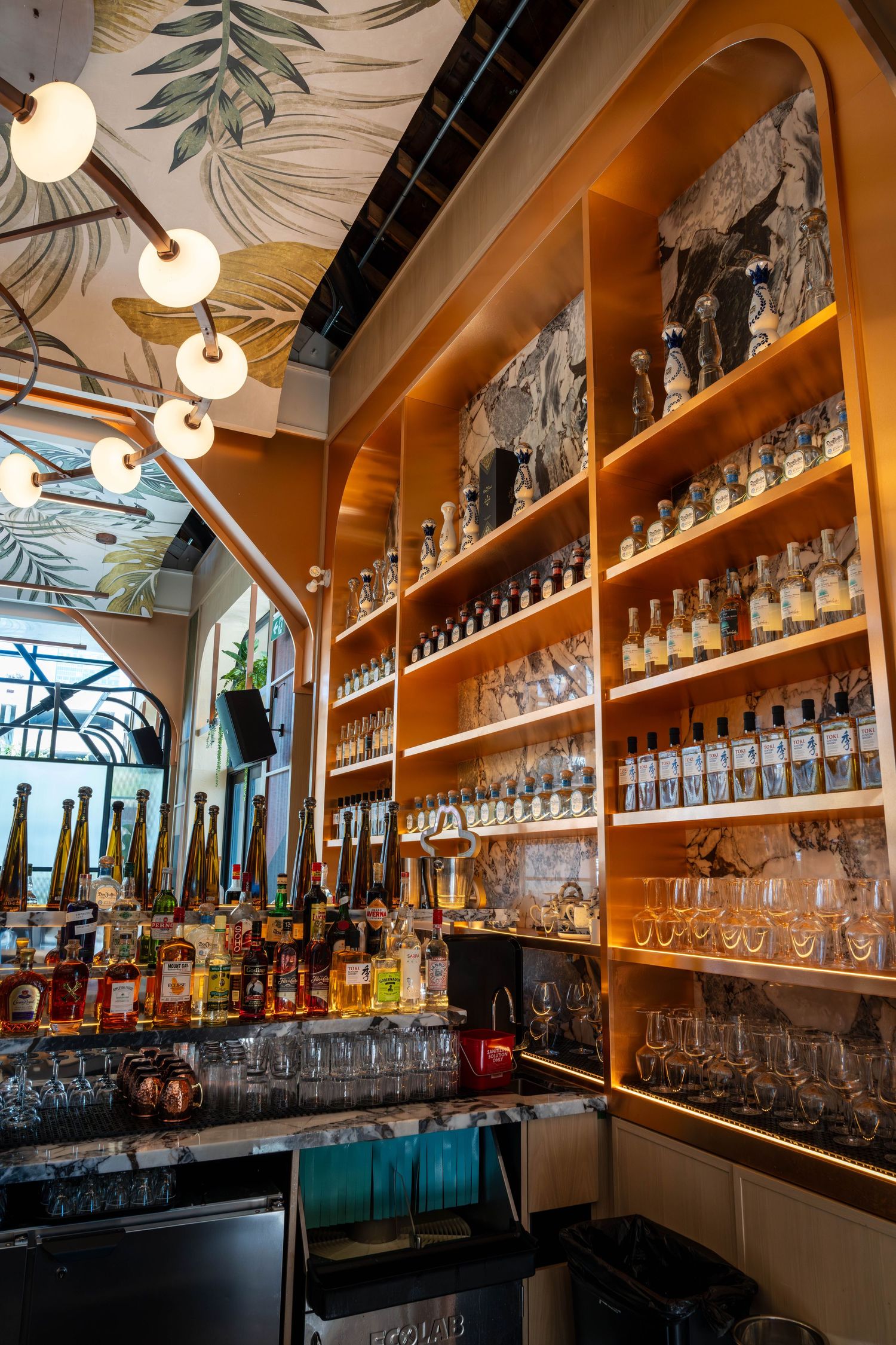 Modern bar interior with wooden shelves displaying glassware and bottles illuminated by warm pendant lighting.