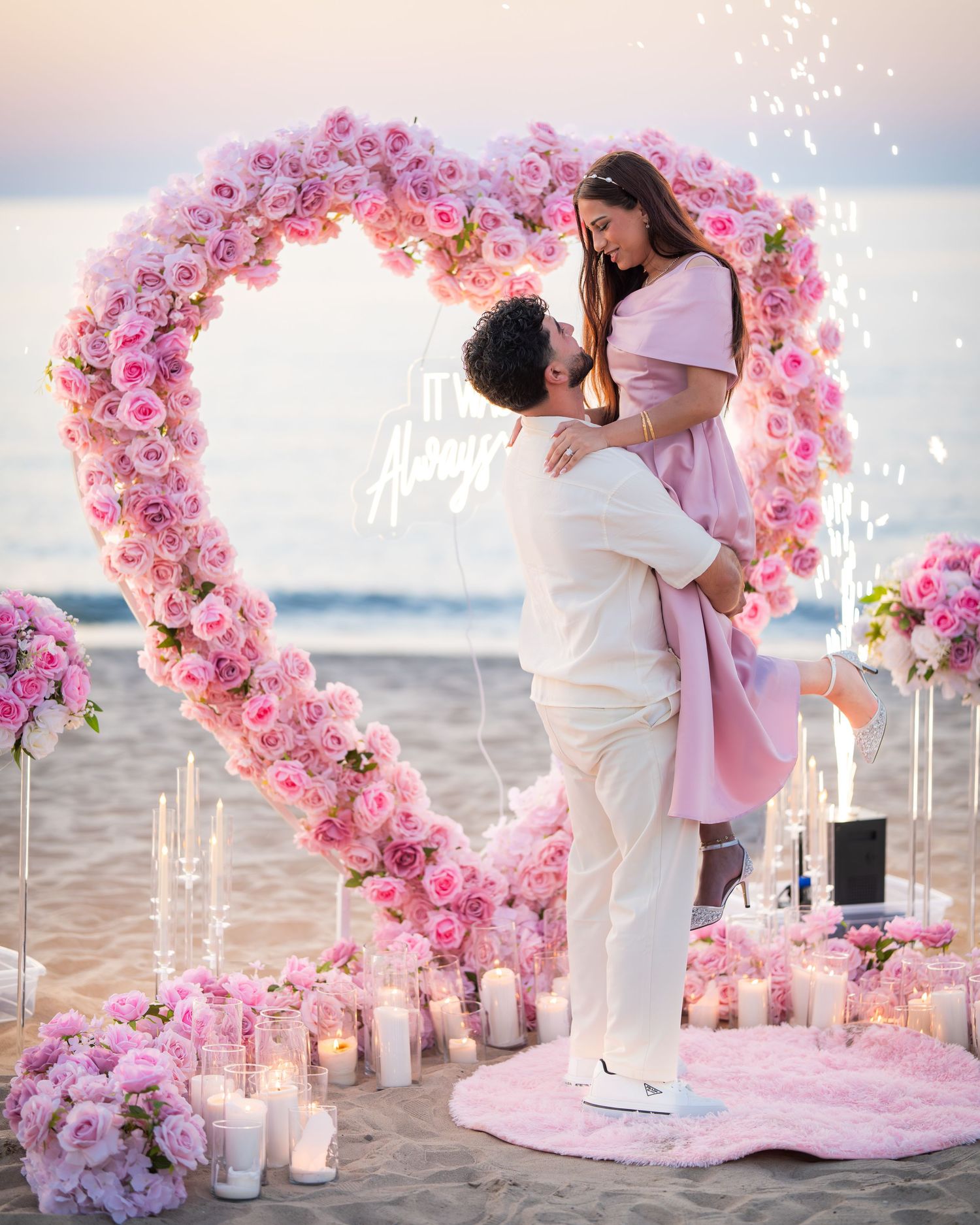 A tender embrace framed by a pink floral heart arch on a beach at sunset.
