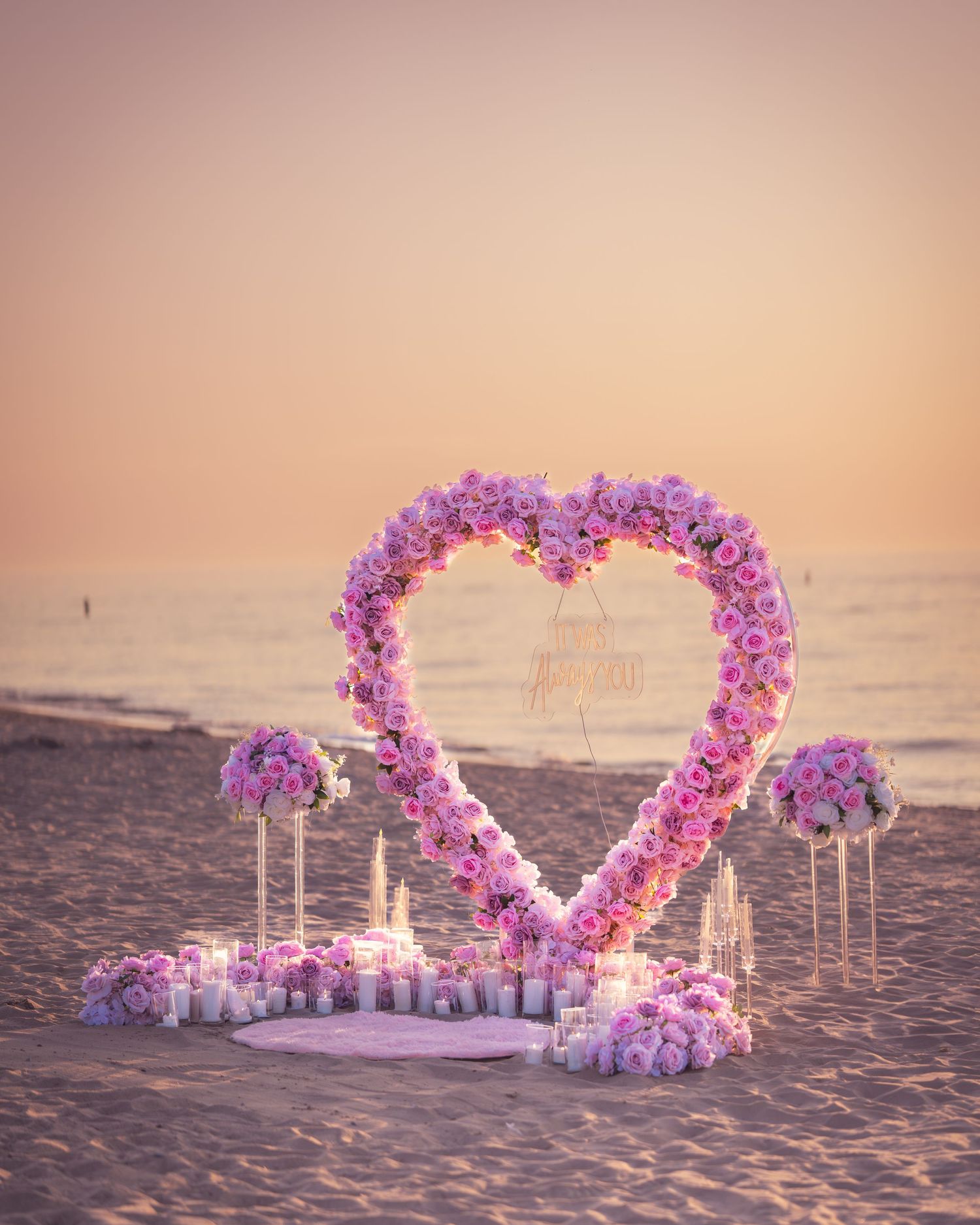 Beach wedding setup with a heart-shaped flower arch and candles glowing in the twilight.