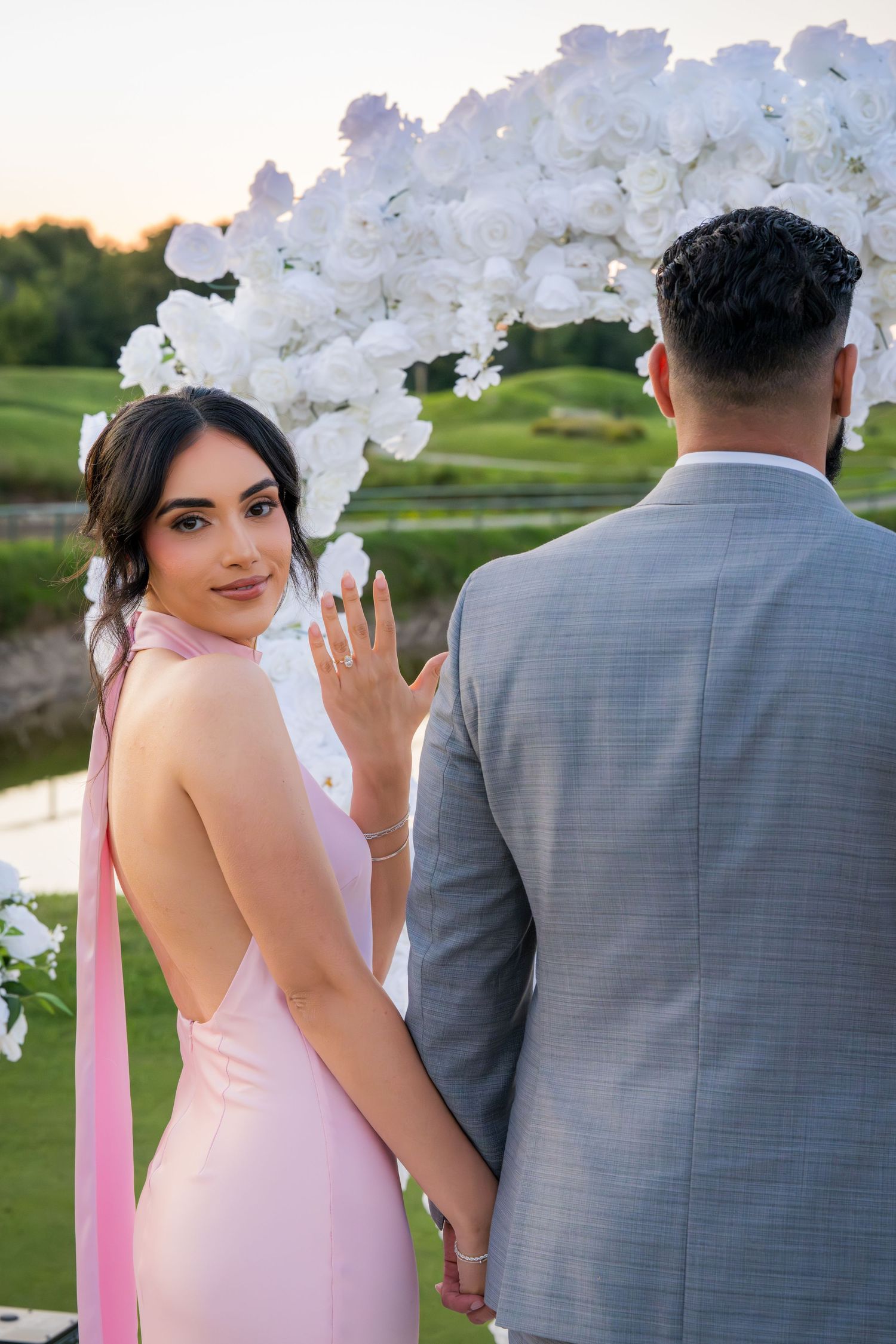 Couple in pink and grey attire pose near white floral arch during sunset.