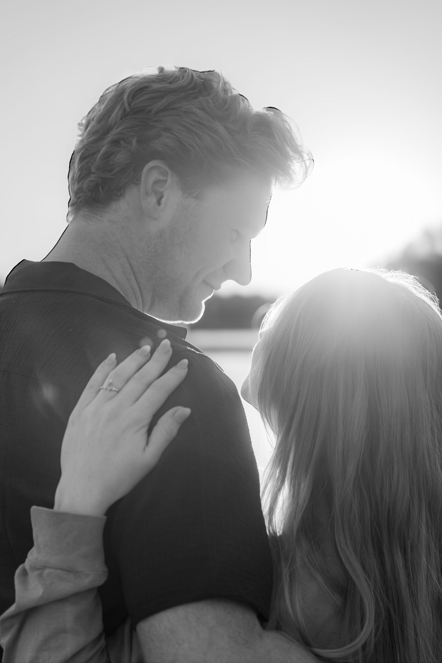 Black and white romantic photo of couple embracing at sunset, woman with engagement ring resting hand on mans chest.