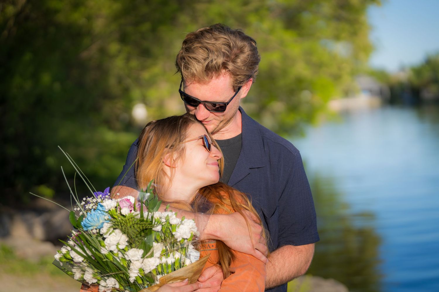 Couple embracing near a lake, woman holding a bouquet of white roses at sunset.