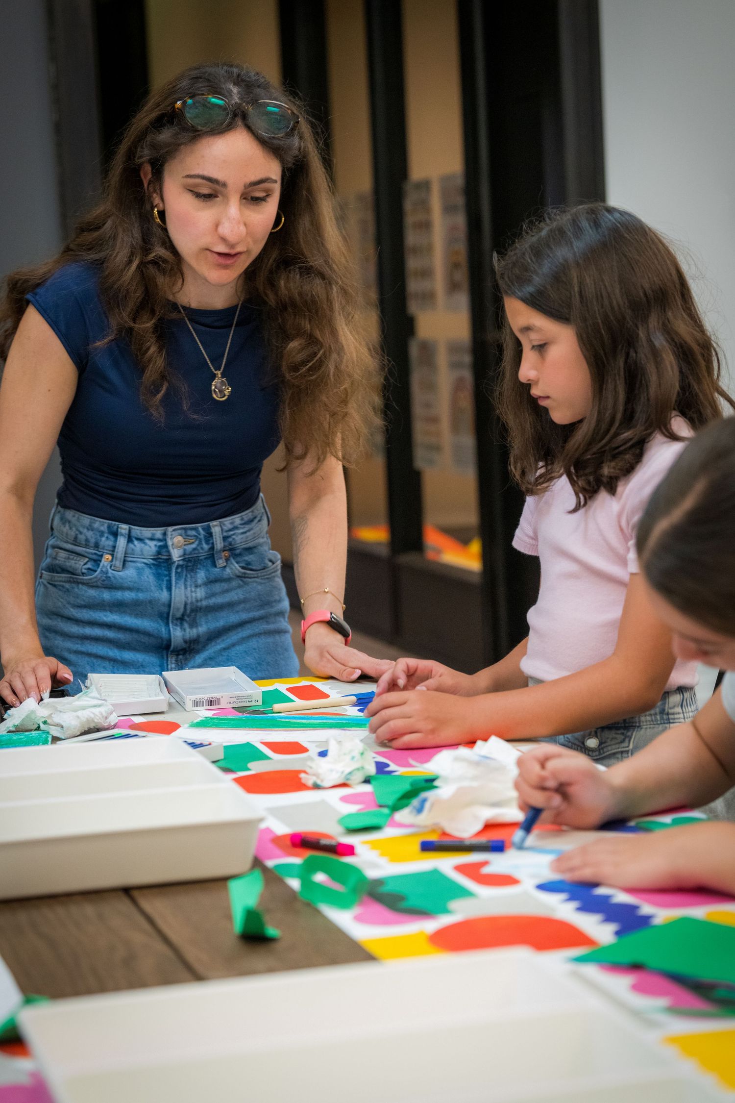 Art teacher with sunglasses helps two young girls arrange colorful cut paper shapes on white canvas.