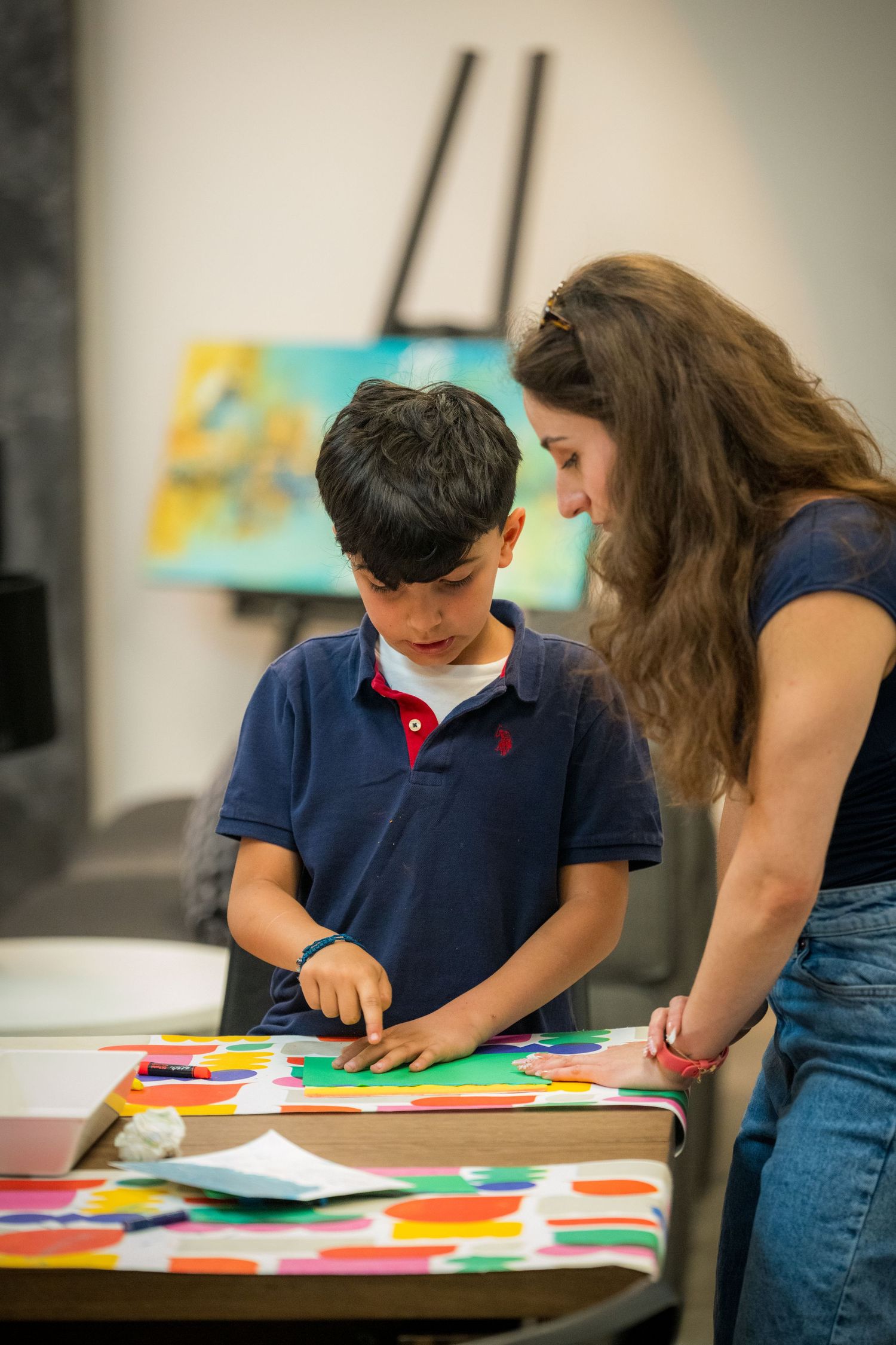 A teacher helps a young boy arrange colorful paper cutouts on a table during an art class.