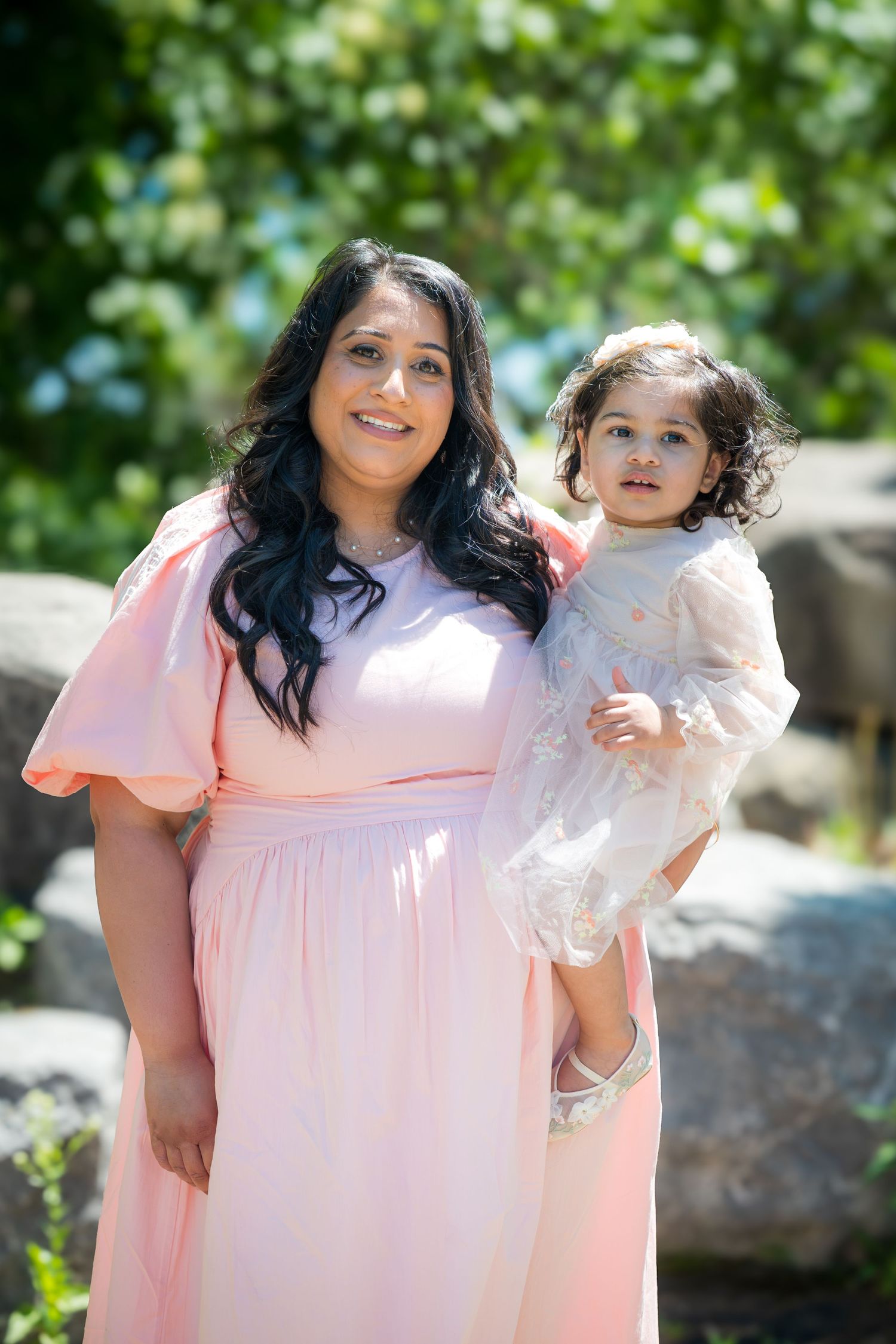 Woman in pink dress holding young child in white outfit, smiling outdoors with green foliage.