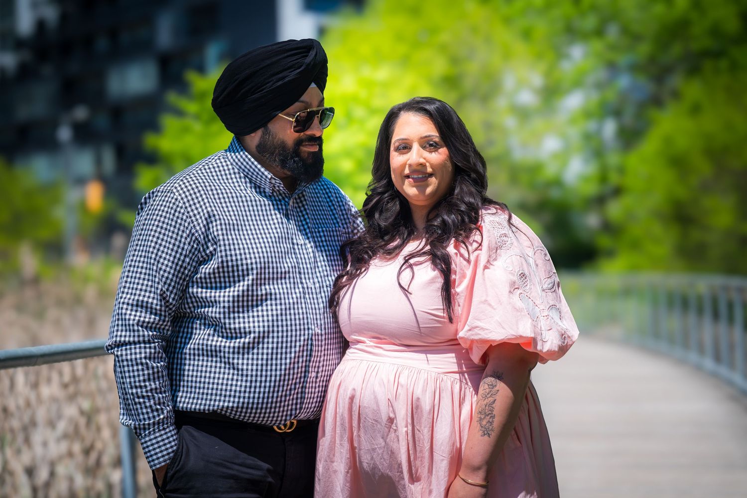 A Sikh man in a turban and a woman in a pink dress smile together on a sunny outdoor walkway.