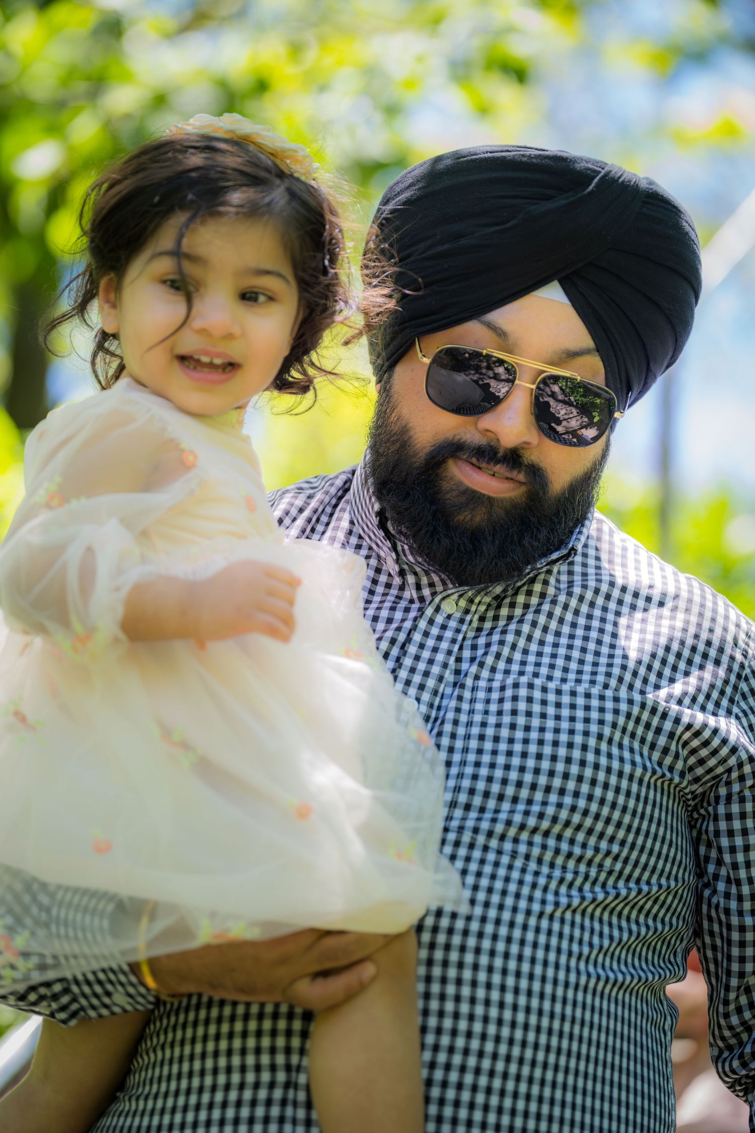 Bearded Sikh father wearing turban and sunglasses holding smiling toddler daughter outdoors.