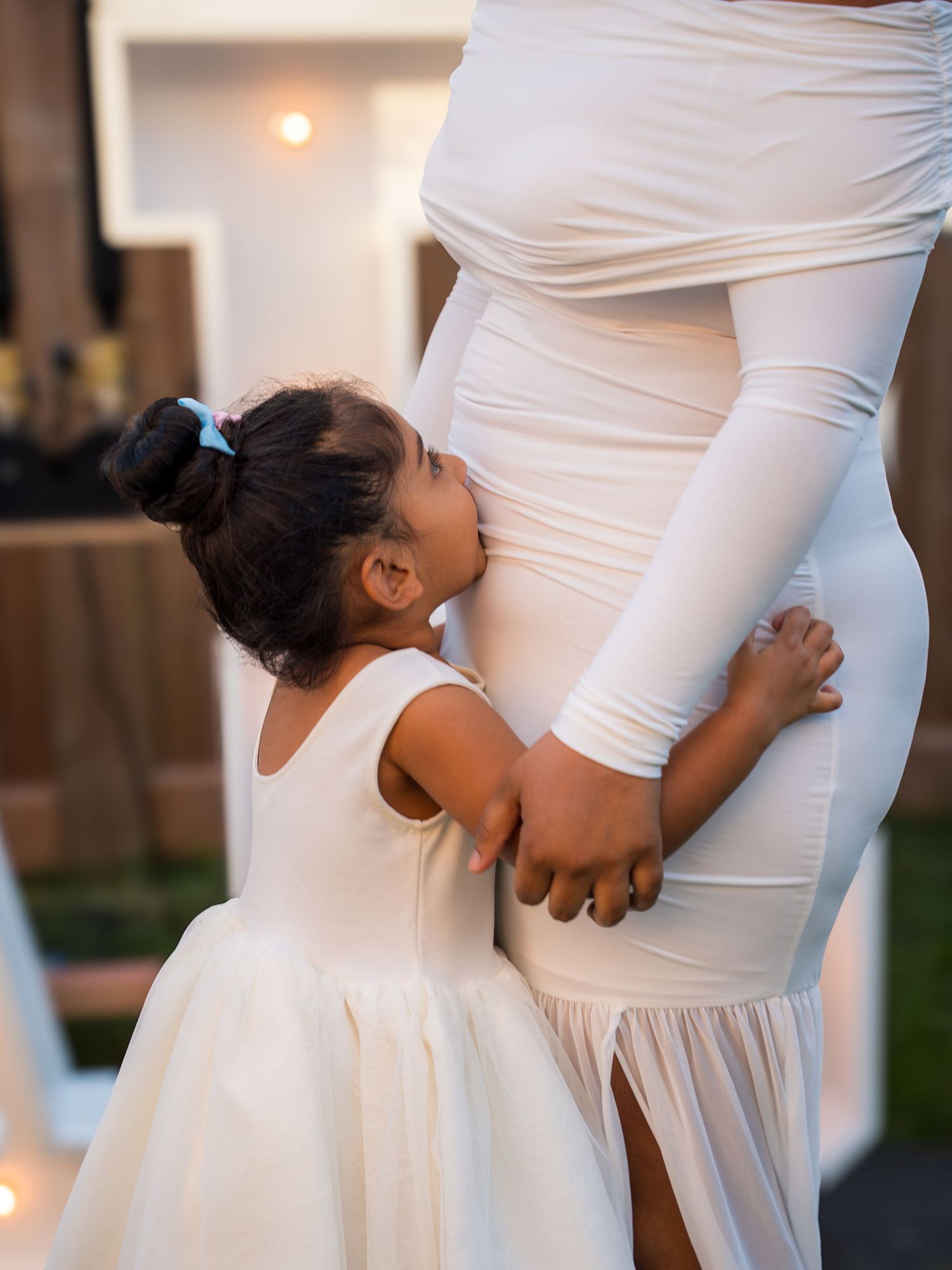 A young girl in a white dress kisses a pregnant woman's baby bump at an outdoor celebration.