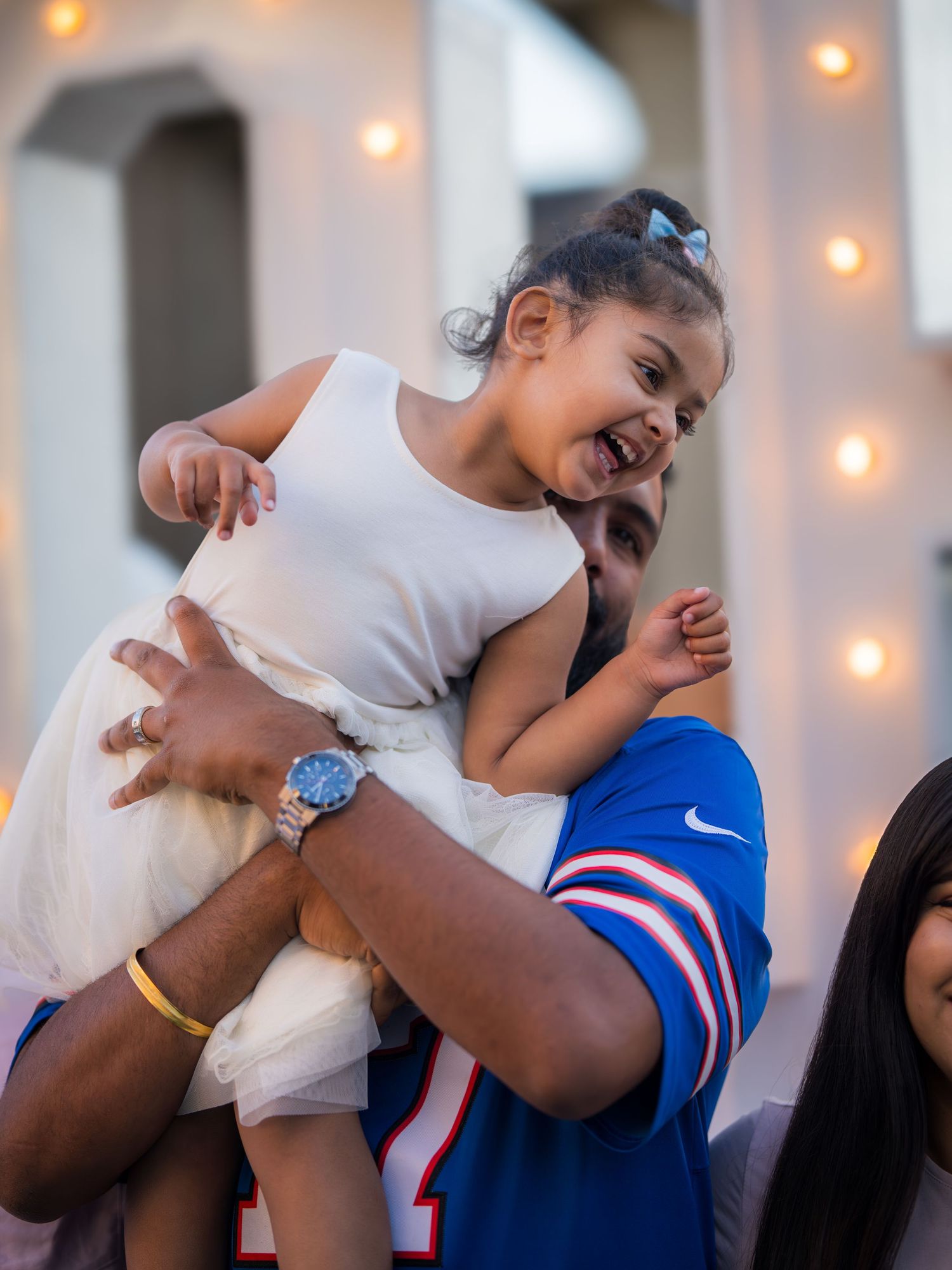 Joyful toddler girl in white dress being lifted up by man in blue Buffalo Bills jersey at outdoor celebration.