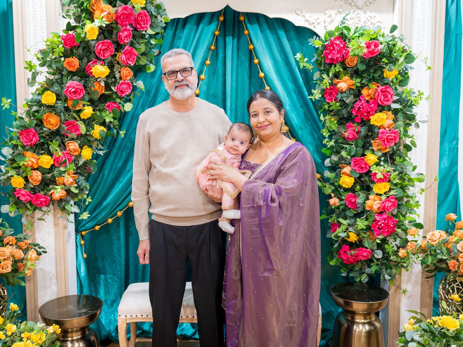 Older couple in beige sweater and purple saree posing with newborn baby at vibrant Indian celebration floral backdrop.
