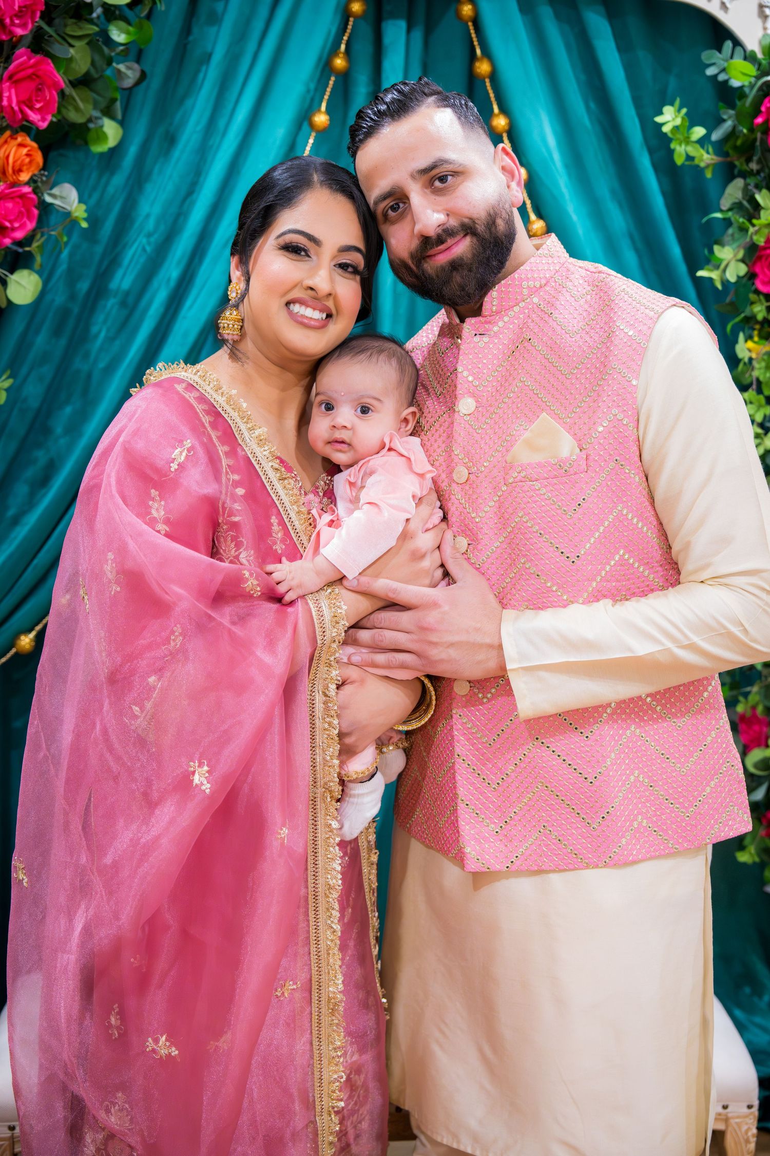 Couple in pink Indian traditional wear smiling with infant at floral decorated ceremony.