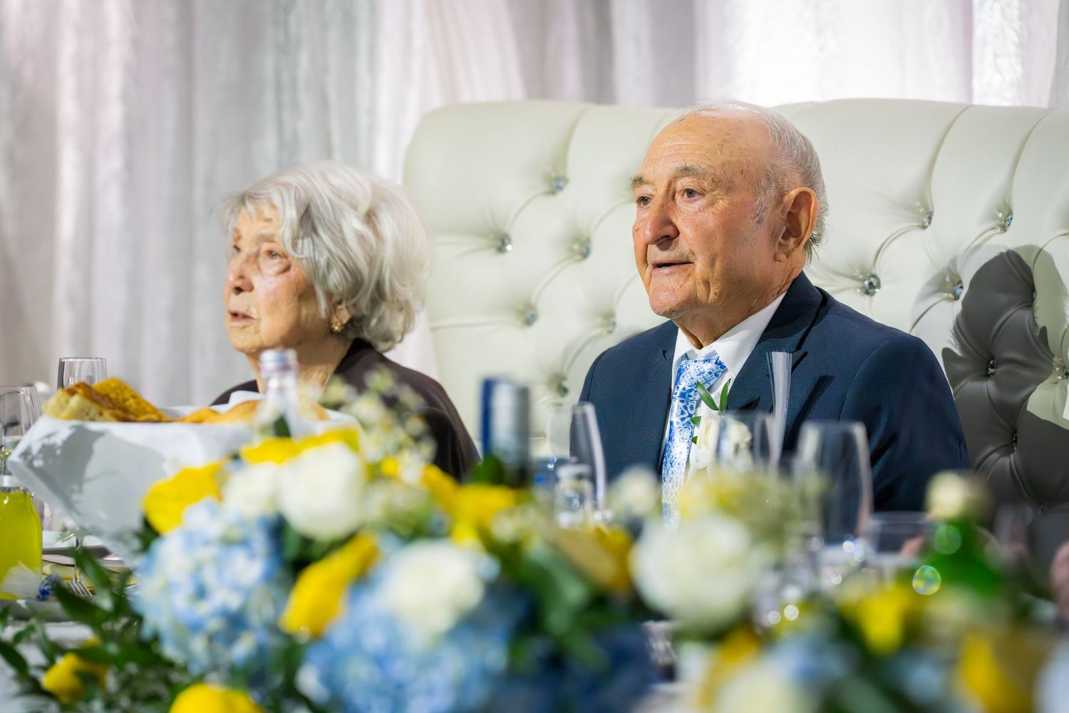 Elderly couple seated at a decorated wedding banquet table with yellow and blue floral centerpieces.