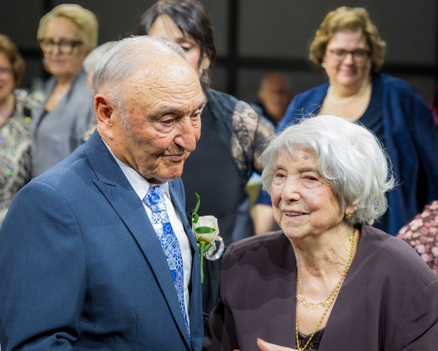 Elderly couple sharing a tender moment at a formal event, man in blue suit with boutonniere, woman in dark top.